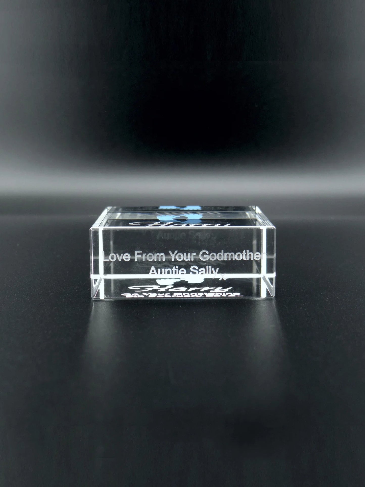 An upright optical crystal plaque that catches and reflects the light. It is engraved with the name "Harry" in a bold, cursive font. Above the name, there are two small, detailed baby footprints coloured in blue. Below the name, the text "On Your Christening 9th February 2014 x" is inscribed in a clear, legible font. The plaque serves as a keepsake to commemorate Harry's christening day. The crystal's transparency and the engraved details are set against a dark background, enhancing their visibility.