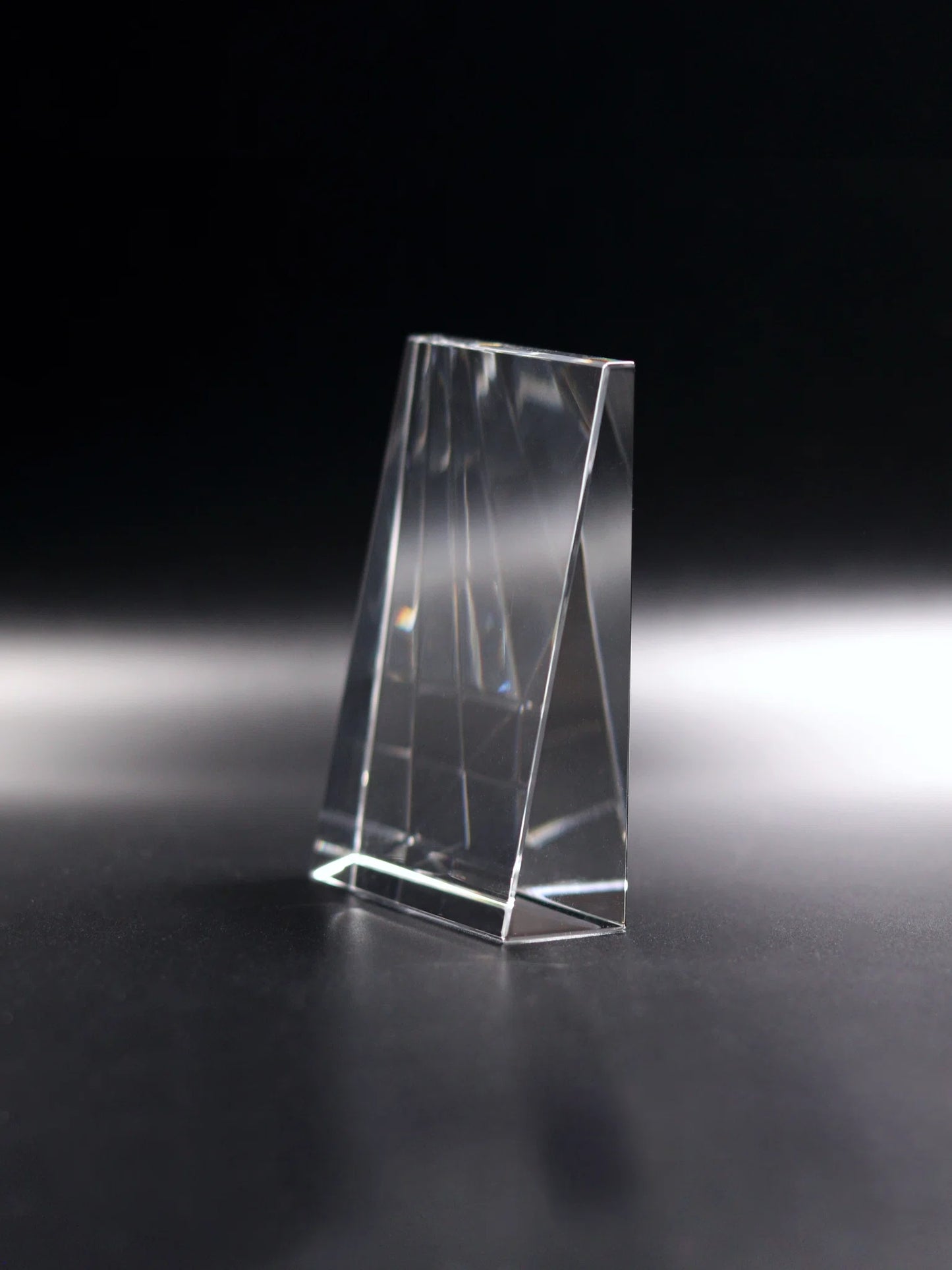 An optical crystal vertical wedge block standing on a reflective dark surface. The block has angled sides that converge to a point at the top, and its transparency allows light to pass through and refract, creating subtle rainbows and a sense of depth within the solid shape. This item is the blank optical crystal ornamental paperweight before being engraved. Its simplicity and clarity make it a versatile piece for various settings.