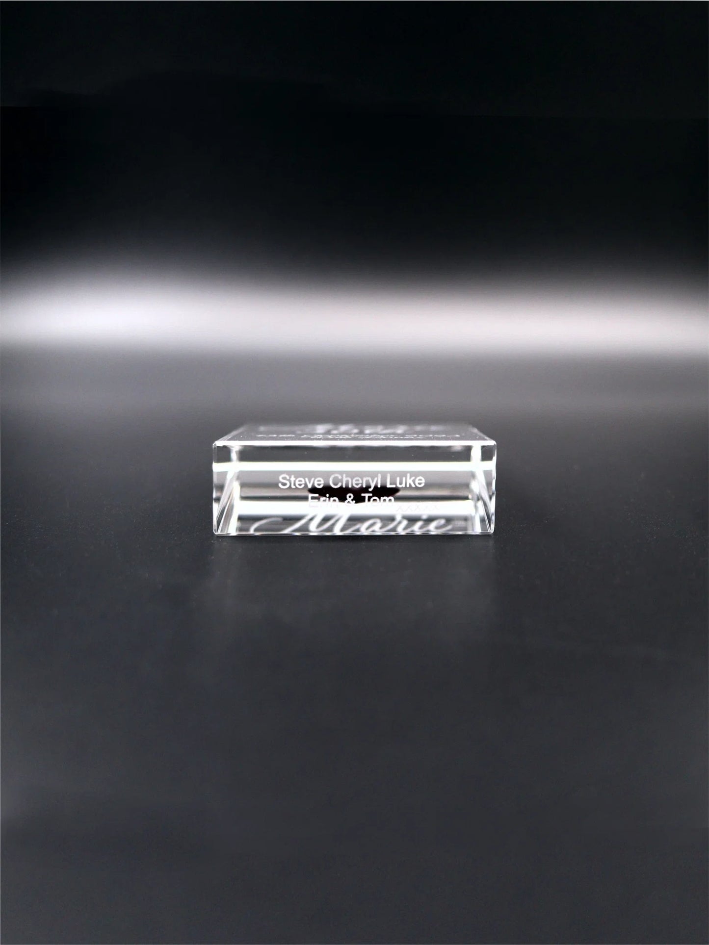 This image showcases the underneath of an optical crystal wedge block placed on a dark, reflective surface. It appears to be engraved with names - "Steve Cheryl Luke Erin & Tom". The clarity of the crystal and the precision of the engraving give the item a delicate and personalized appeal, potentially serving as a memento or gift.