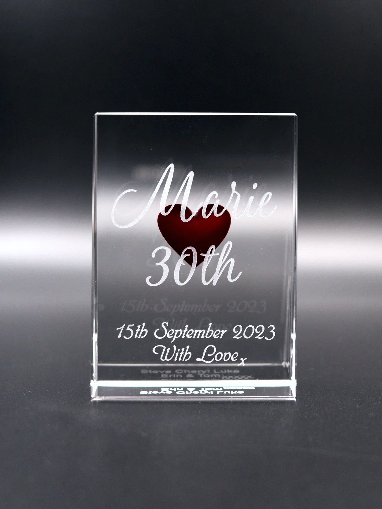 An optical crystal vertical wedge standing upright on a reflective surface. It is elegantly engraved with "Marie 30th" in script font, celebrating a milestone. Below, "15th September 2023 With Love" is inscribed, suggesting the date of the occasion. A red heart is deeply engraved on the reverse side, adding a pop of colour and sentiment to the commemorative piece. The sleek design and personalized engraving make it an ideal keepsake for marking special moments.