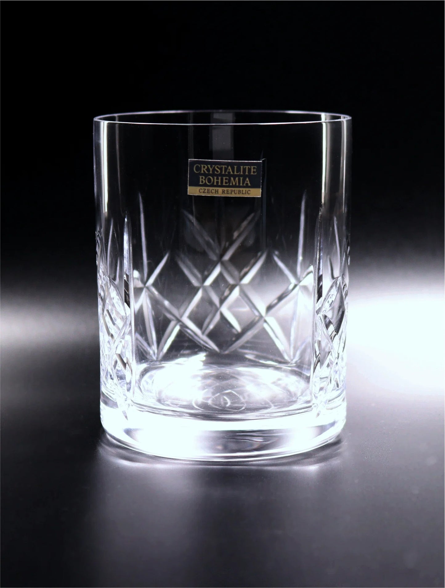 A pristine, cylindrical crystal glass from Crystalite Bohemia, featuring a textured lower half with meticulously cut diamond patterns that catch and refract light, enhancing its sparkling clarity. The smooth upper half is unadorned, leading to a thin rim, and showcases a label affirming its Czech Republic origin. The whisky glass is panelled and awaits personalization or can stand alone as a testament to fine craftsmanship.