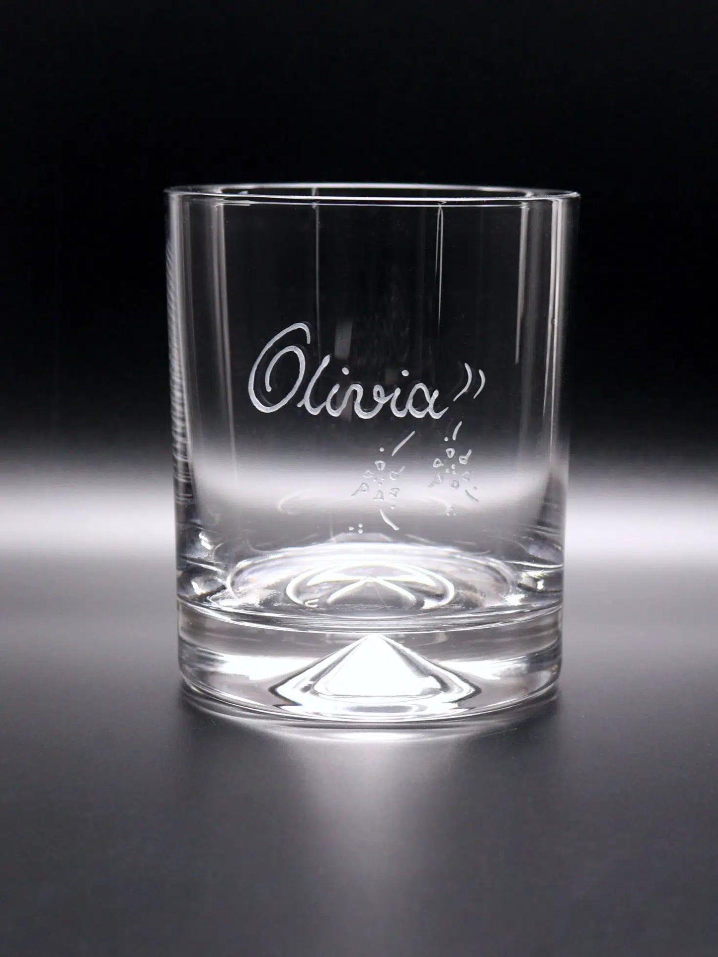 This image shows a clear, cone-based whisky glass with a sturdy bottom and smooth, straight sides. It features the name "Olivia" elegantly hand-engraved on the front in a flowing script font, accompanied by decorative elements that suggest a personal touch. The glass is set against a dark background, highlighting its clarity and the fine detail of the engraving. This glass combines functionality with a personalized aspect, intended as a special gift or keepsake.