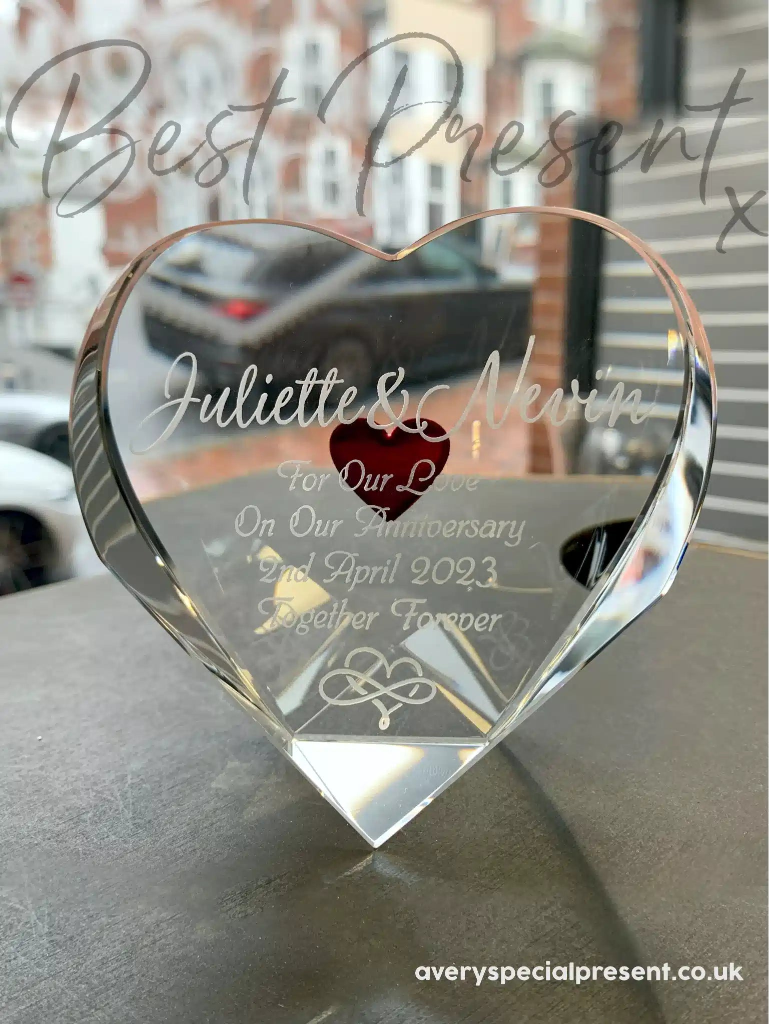 Triple Heart, an Optical Crystal Heart, Deeply Engraved Red Heart on the reverse, engraved with "Juliette & Nevin For Our Love On Our Anniversary 2nd April 2023 Together Forever" along with an eternity and heart symbol, displayed looking out of a shop window onto the high street backdrop, signifying a timeless romantic gift.