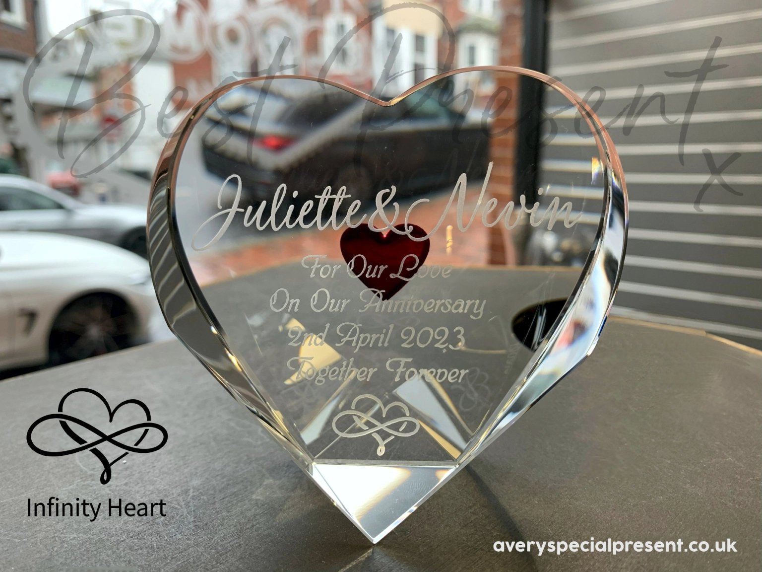 An Optical Crystal Heart, Deeply Engraved Red Heart on the reverse, engraved with "Juliette & Nevin For Our Love On Our Anniversary 2nd April 2023 Together Forever" along with an eternity symbol, displayed looking out of a shop window onto the high street backdrop, signifying a timeless romantic gift. With text overlay ‘Best Present x’ and an infinity and heart design titled ‘Infinity Heart’.