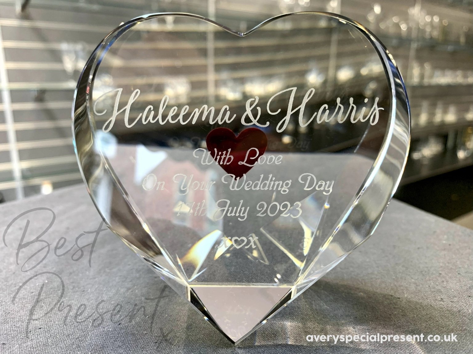An optical heart-shaped crystal keepsake with a red heart engraved on the reverse and "Haleema & Harris With Love On Your Wedding Day 4th July 2023" and a kiss heart kiss engraved on the front, displayed in interior shop setting, creating a reflective and romantic memento.