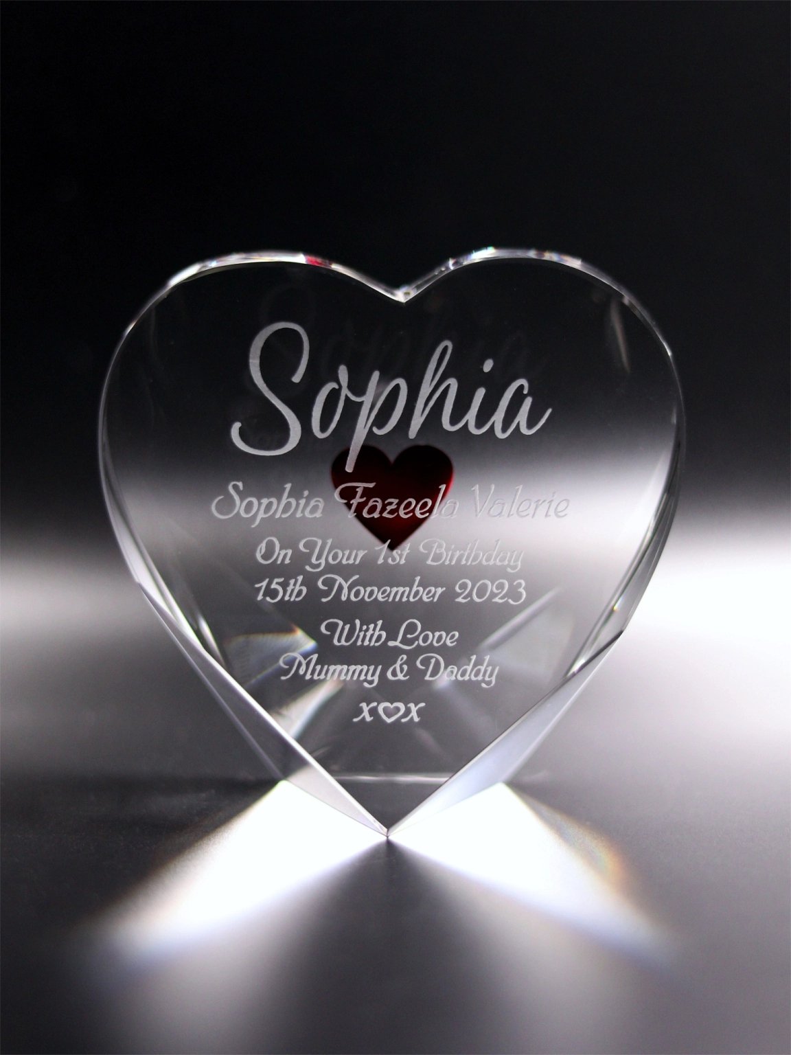 An optical crystal heart block with a red heart engraved on the reverse and "Sophia” as a focal name, and “Sophia Fazeela Valerie On Your 1st Birthday 15th November 2023 With Love Mummy & Daddy x ♡ x" on the front, set against a dark backdrop that highlights the clarity and personalized details of the keepsake.