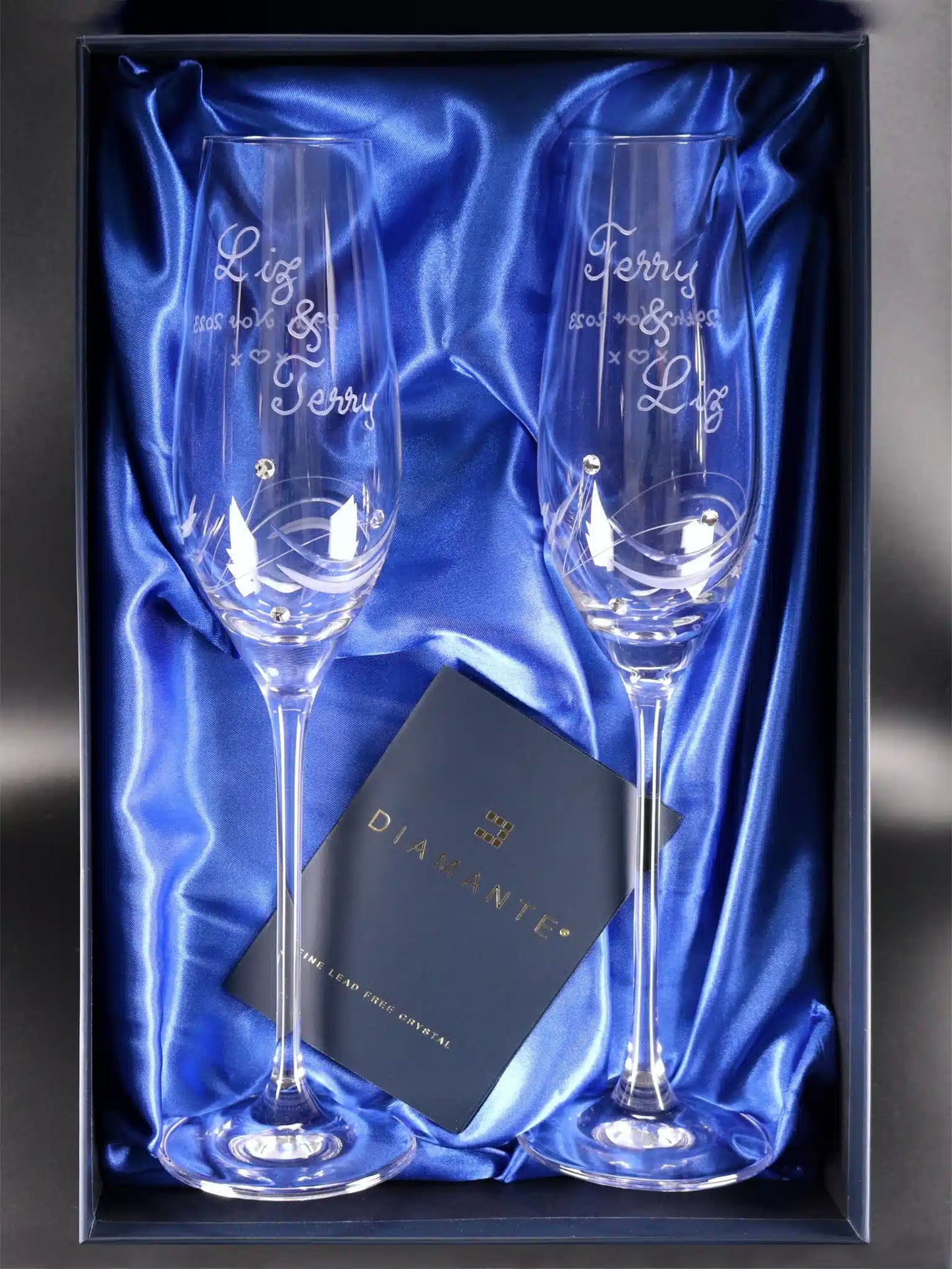 Two elegant Diamante champagne flutes with precision swish cut detailing and Swarovski crystals, each glass engraved with names "Liz & Terry," reversed on the other, and a commemorative date "29th Nov 2023" on one side, with a playful "kiss, heart, kiss" pattern on the reverse. Presented in a luxuriousblue satin-lined box, signifying a premium gift choice.