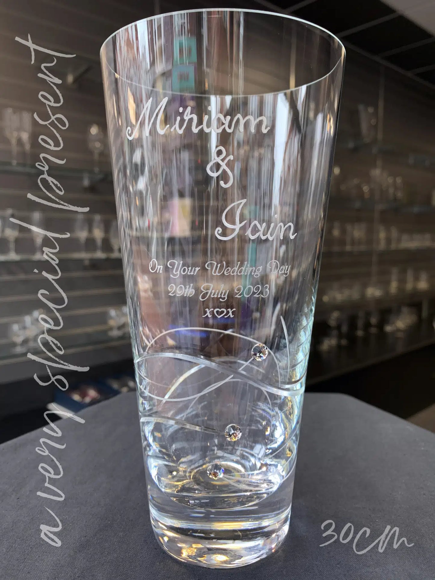 A tall, hand-engraved vase with the names "Miriam & Iain" etched elegantly above a sandblasted message "On Your Wedding Day 29th July 2023" and an elegant "kiss, heart, kiss" with swirling cut glass and three sparkling crystals embedded into the design. The vase stands on a grey surface with the backdrop of a shop wall with glass shelves with glassware. Overlayed text "30cm" indicating its height, showcasing its stately presence.