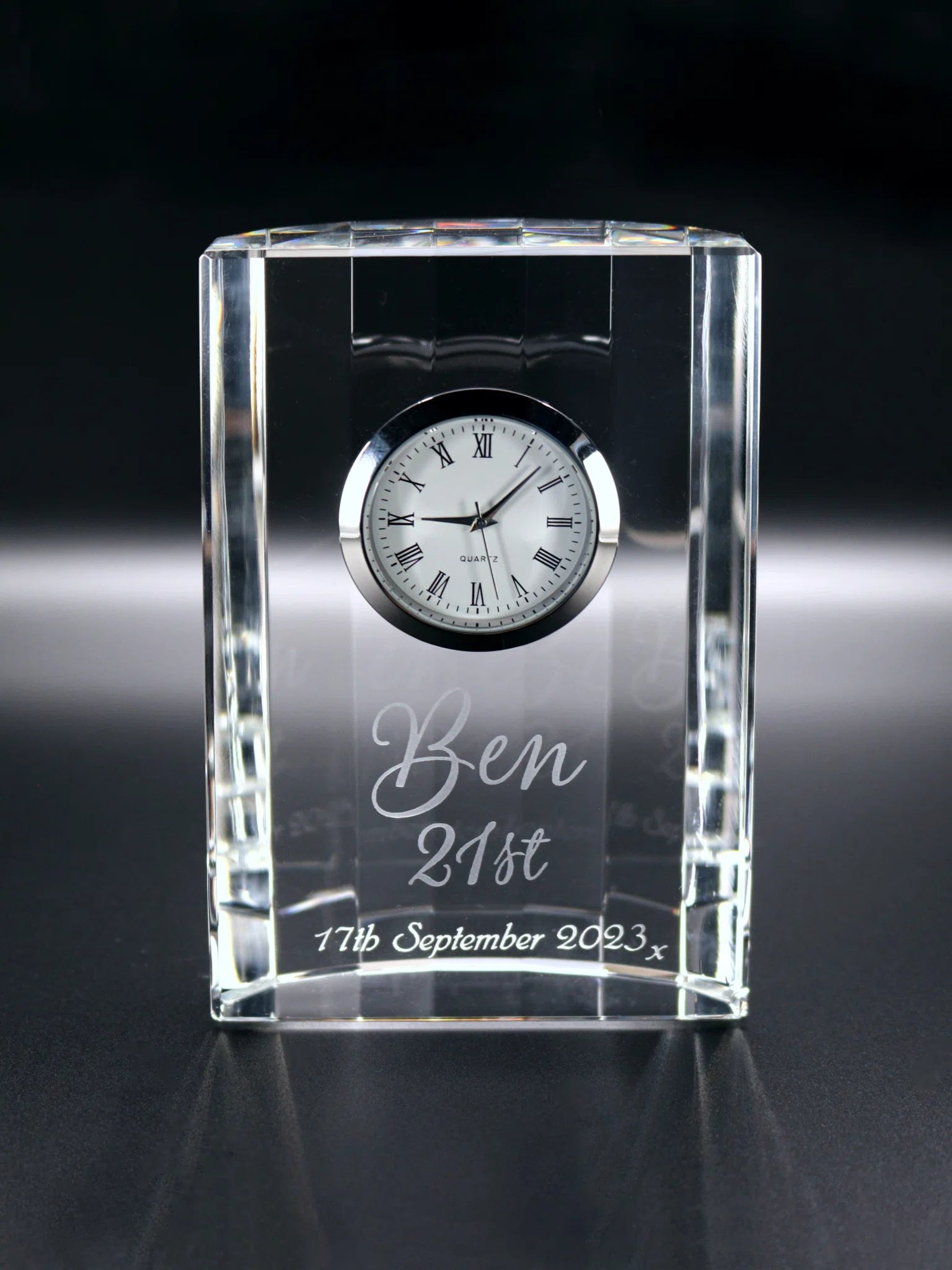 A rectangular optical crystal clock with a series of facets forming a scalloped back the crystal clock stands elegantly with a built-in timepiece at the upper half. The clock face is classic, featuring roman numerals and a quartz movement. Below the clock, the glass is artfully engraved with "Ben 21st" in a stylish, cursive font, followed by "11th September 2023," marking a significant date, for a 21st birthday. The glass's edges catch the light, showcasing the clarity and quality of the material.