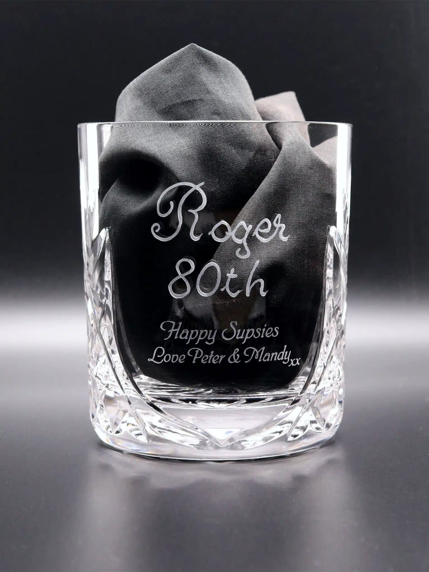 A 24% lead crystal whisky glass with a diamond-cut base, hand engraved and sandblasted with "Roger 80th Happy Surspies Love Peter & Mandy xx". The glass cradles a folded black napkin, set against a dark background, highlighting the personalized message and intricate design details.