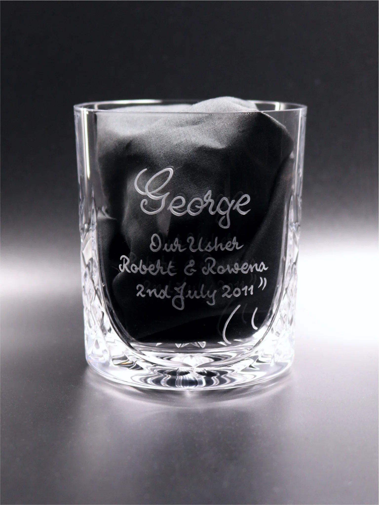 A 24% lead crystal whisky glass with a detailed diamond pattern on the base, engraved with the name "George" and the title "Our Usher" followed by "Robert & Rowena 2nd July 2011" finished with an elegant swish. The glass contains a black fabric napkin, set against a reflective dark surface, emphasizing the glass's personalized engraving and craftsmanship.