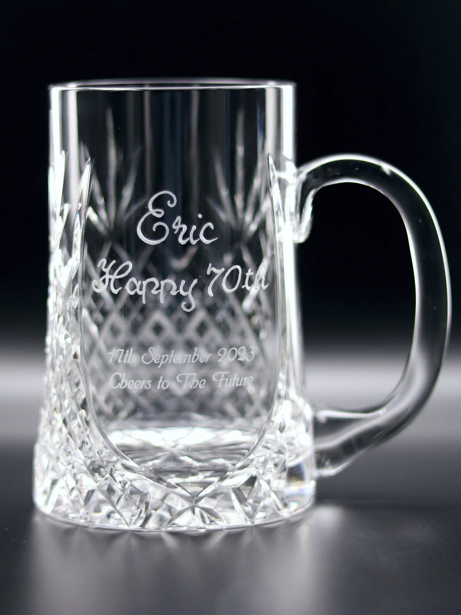 A 24% crystal pint tankard with a diamond-cut pattern on the lower half, featuring a personalized engraving that reads "Eric Happy 70th" hand engraved and “11th September 2023 Cheers to The Future" crisply sandblasted. The clear, intricate design and the heartfelt inscription celebrate a milestone with elegance and a toast to the years ahead.