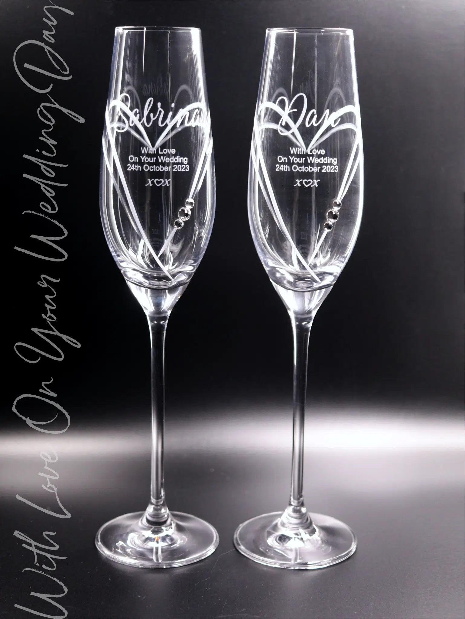 A pair of personalised Diamante champagne flutes with names "Sabrina" and "Dan" engraved in a flowing sandblasted script, with a Heart cut into the glass framing the engraving. Additional engraving reads "With Love On Your Wedding 24th October 2023 x♡x"