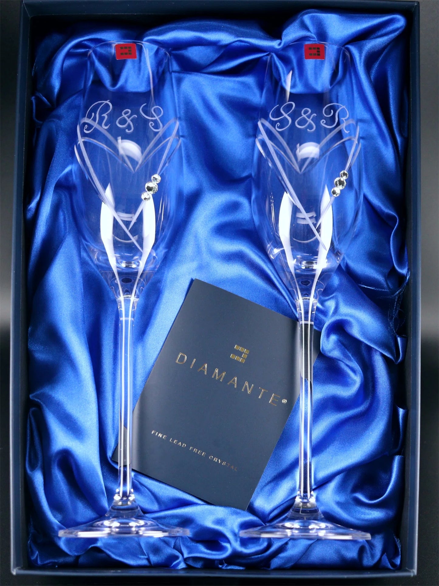 Two Diamante champagne flutes with heart-shaped cutting patterns and three Swarovski crystals, displayed in a satin-lined box. The glasses features the monogram "R&S" and “S&R” engraved on each in an elegant hand engraved script. The flutes have slender stems and are part of a lead-free crystal collection. A dark blue card with gold text reads "DIAMANTE - FINE LEAD FREE CRYSTAL."