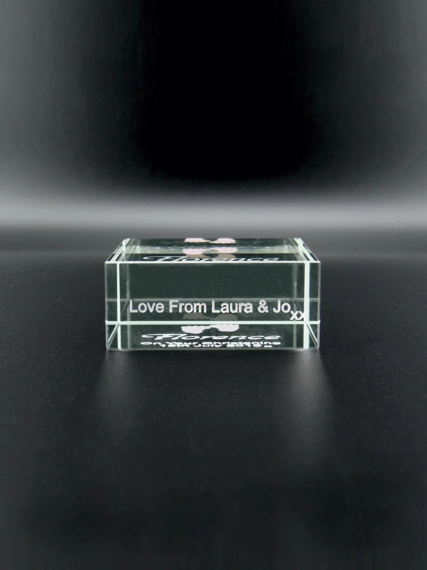 This image shows the underneath of a jade glass block placed on a dark surface. The block is engraved with "Love From Laura & Jo xx" in a simple, elegant script. The transparency of the glass and the reflective quality of the surface create a sense of depth and luminosity, making the text appear to float within the block. It seems to be a personal token of affection from two individuals named Laura and Jo.