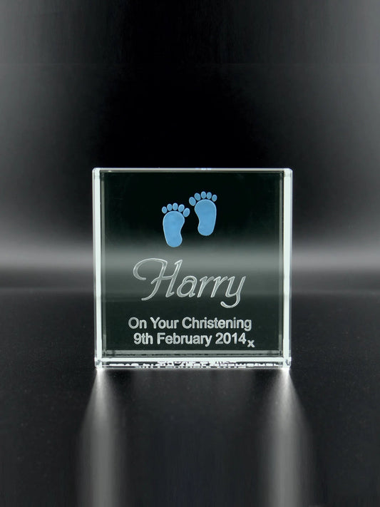 5cm square jade glass plaque with smooth edges, standing upright. It features a deep-etched engraving of two baby footprints at the top, tinted with a subtle blue shade. Below the footprints, the name "Harry" is engraved in a cursive, elegant font. A commemorative inscription reads "On Your Christening 9th February 2014," engraved in a clear, legible script. The glass has a glossy finish, reflecting the light and enhancing the three-dimensional quality of the engravings.