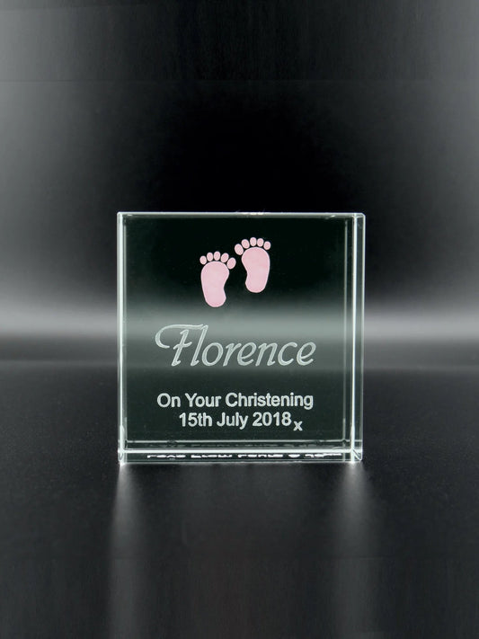 This image features a jade glass plaque that stands upright on a dark surface. It's engraved with the name "Florence" in a cursive font, followed by "On Your Christening 15th July 2018" in a clearer, block-type font. At the top of the plaque, there are two small, pink baby footprints, suggesting the plaque is a commemorative item for a child's christening day. The smooth edges of the plaque reflect the light, giving it a delicate luminosity.