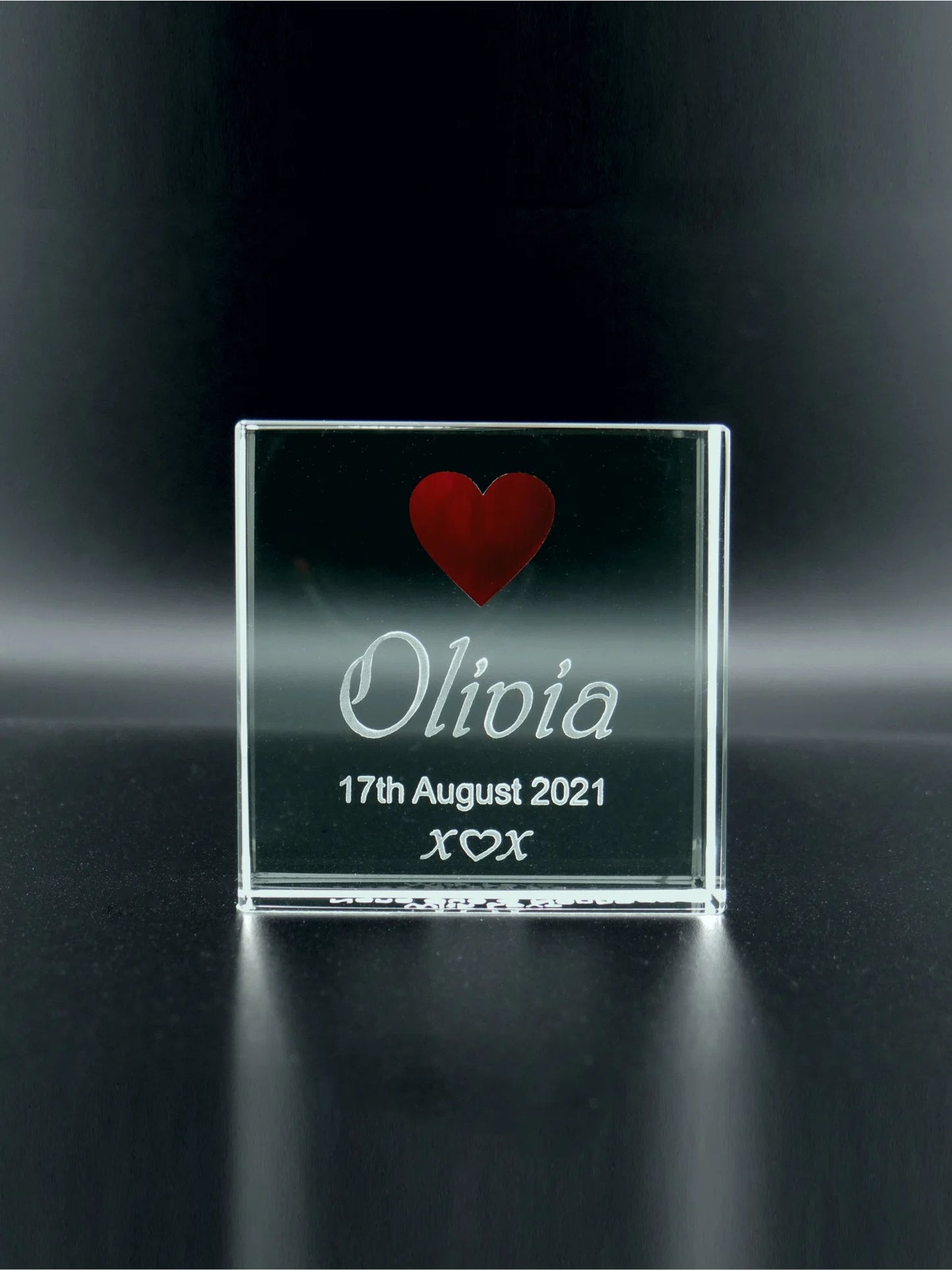 A jade glass square plaque. It's engraved with the name "Olivia" in a flowing, cursive script. Below the name, the date "17th August 2021" is marked, and beneath that, three small kisses represented as "x ♡ x". Above the name, a solid red heart adds a pop of colour and emotion to the piece. The plaque itself stands on a reflective surface, casting a subtle glow and giving the etchings a floating appearance. This item likely signifies a sentimental occasion on the specified date.