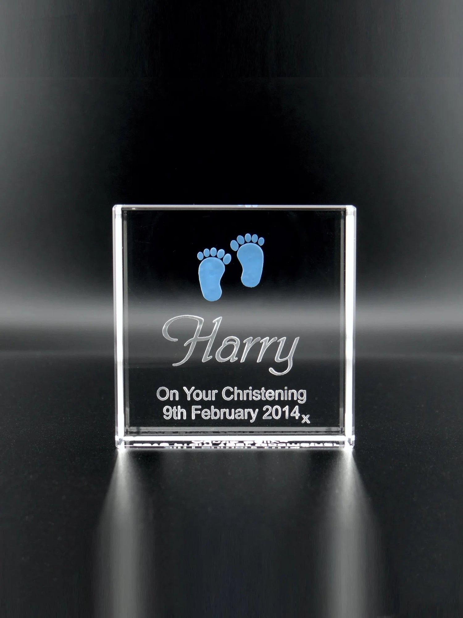 An upright optical crystal plaque that catches and reflects the light. It is engraved with the name "Harry" in a bold, cursive font. Above the name, there are two small, detailed baby footprints coloured in blue. Below the name, the text "On Your Christening 9th February 2014 x" is inscribed in a clear, legible font. The plaque serves as a keepsake to commemorate Harry's christening day. The crystal's transparency and the engraved details are set against a dark background, enhancing their visibility.