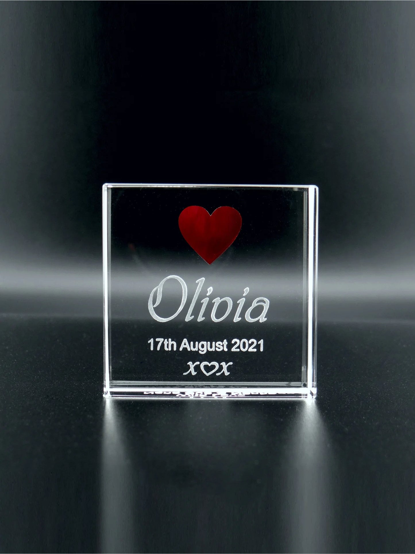 An optical crystal plaque that stands upright against a dark background. It features a red heart at the top, followed by the name "Olivia" written in cursive script. Below the name, the date "17th August 2021" is engraved, accompanied by three kisses represented as "x ♡ x". The simplicity of the design—the bold red heart above the graceful lettering—gives the plaque a charming and sentimental quality. The reflective surface adds depth and a hint of luminosity to the keepsake.