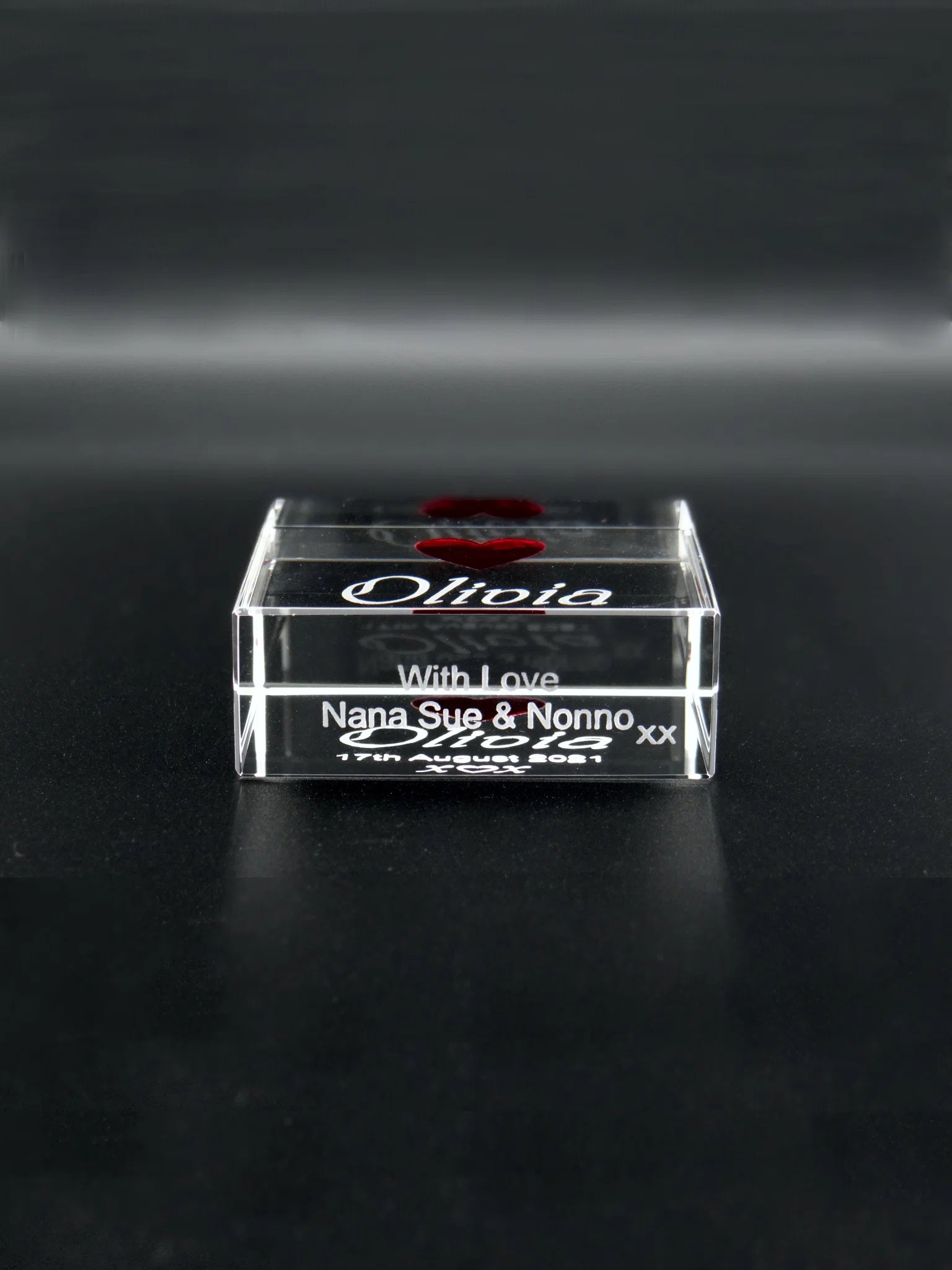 The image displays the underneath of an optical crystal block on a dark background, which reflects light along its polished edges. Engraved within the crystal is the name "Olivia" in an elegant cursive script, accentuated by a red heart above the name. Beneath the block reads "With love Nana Sue & Nonno xx," indicating the block is a sentimental gift from grandparents. The piece has a personal and affectionate feel, likely serving as a cherished keepsake.