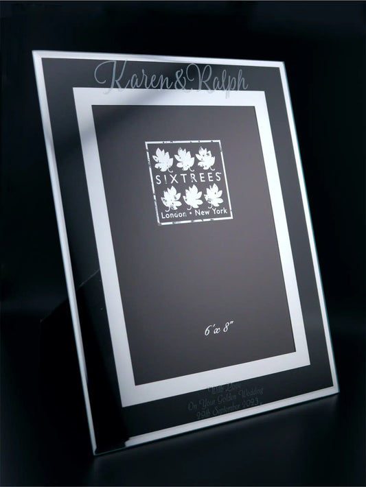 A sleek, clear glass photo frame with a mirrored border standing on a reflective surface. The frame is elegantly engraved with "Karen & Ralph" at the top and "With Love On Your Golden Wedding 29th September 2023" at the bottom. Centred within the border is the branding "SIXTREES London • New York" and the photo area "6 x 8". The frame's sophisticated design is both modern and timeless, suitable for celebrating a significant marital milestone.