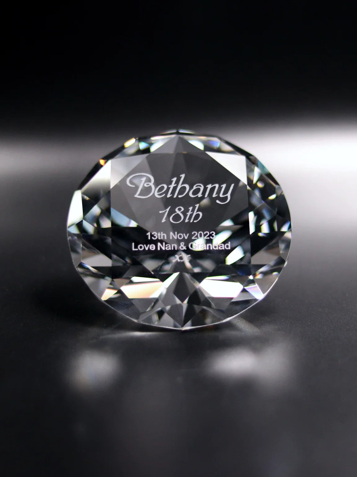 The image shows a faceted optical crystal with a round cut, resembling a large gemstone, resting on a dark surface. It is engraved with the name "Bethany 18th" in elegant script, followed by "13th Nov 2023" and "Love Nan & Grandad" below. The crystal's many facets catch the light, creating a sparkling effect that enhances the visibility of the engraving, suggesting a commemorative gift for Bethany's 18th birthday.