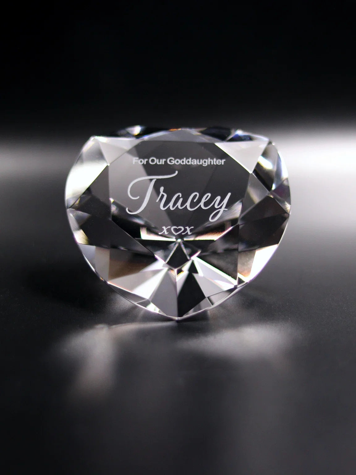 The image features a heart-shaped optical crystal with faceted edges, creating a reflective, sparkling appearance on a dark background. It's engraved with "For Our Goddaughter Tracey x ♡ x", signifying a heartfelt message intended as a gift. The facets catch and refract light, giving the crystal depth and brilliance, while the engraved message adds a personal touch.