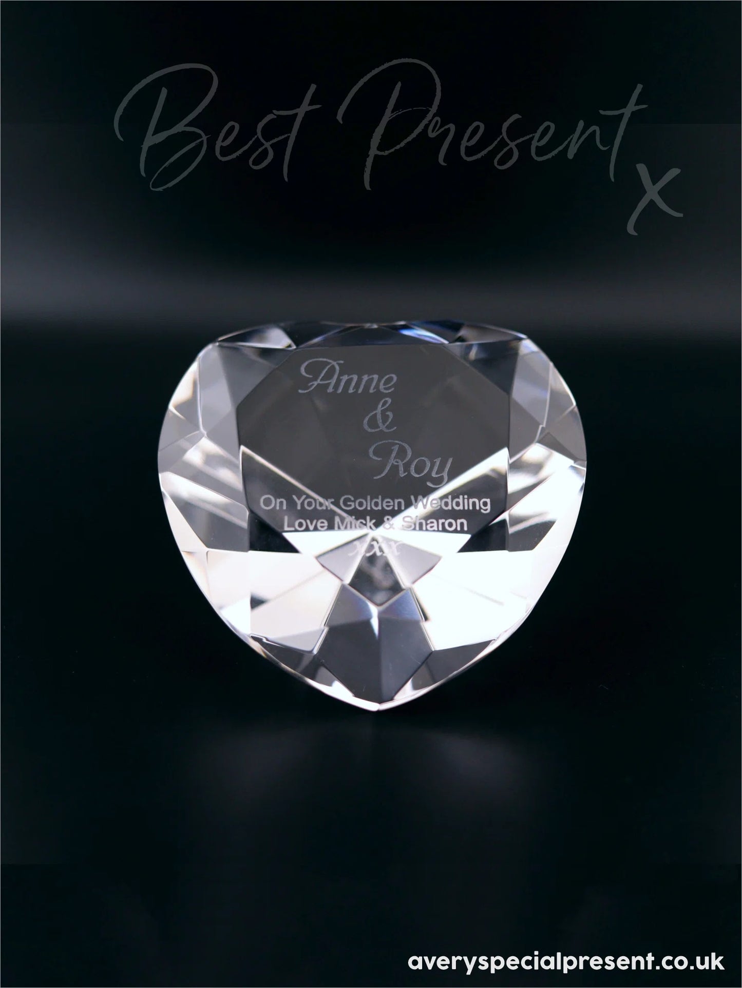 The image features a heart-shaped optical crystal with faceted edges, creating a reflective, sparkling appearance on a dark background. It's engraved with "Anne & Roy On Your Golden Wedding Love Mick & Sharon xxx", signifying a heartfelt message intended as a gift. The facets catch and refract light, giving the crystal depth and brilliance, while the engraved message adds a personal touch.