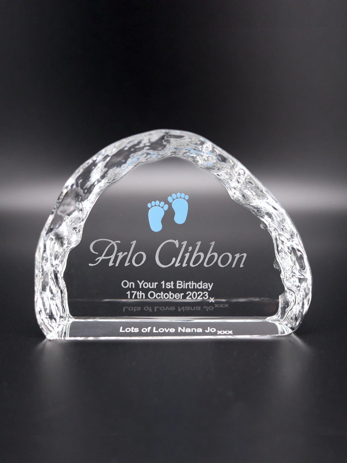 An arched, optical crystal ornament with textured edges that mimic the roughness of ice. Engraved within the arch are the words "Arlo Clibbon On Your 1st Birthday 17th October 2023 x", with two small blue baby footprints above, adding a charming detail. Inscribed on the base visible from the front "Lots of Love Nana Jo xxx". The ornament is placed against a dark background, which highlights the clarity of the crystal and the detail of the engravings, making it a special keepsake