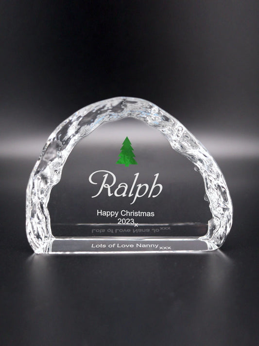 An arched crystal ornament with ice-like texture on its edges. It is engraved with "Ralph Happy Christmas 2023 x", underneath is visible from the front "Lots of Love Nanny xxx". At the center, just above the name Ralph, is a small green Christmas tree, adding a festive touch to the piece. The ornament stands against a dark background, which contrasts with the clear crystal, highlighting the engravings and the tree, making it a special holiday keepsake.