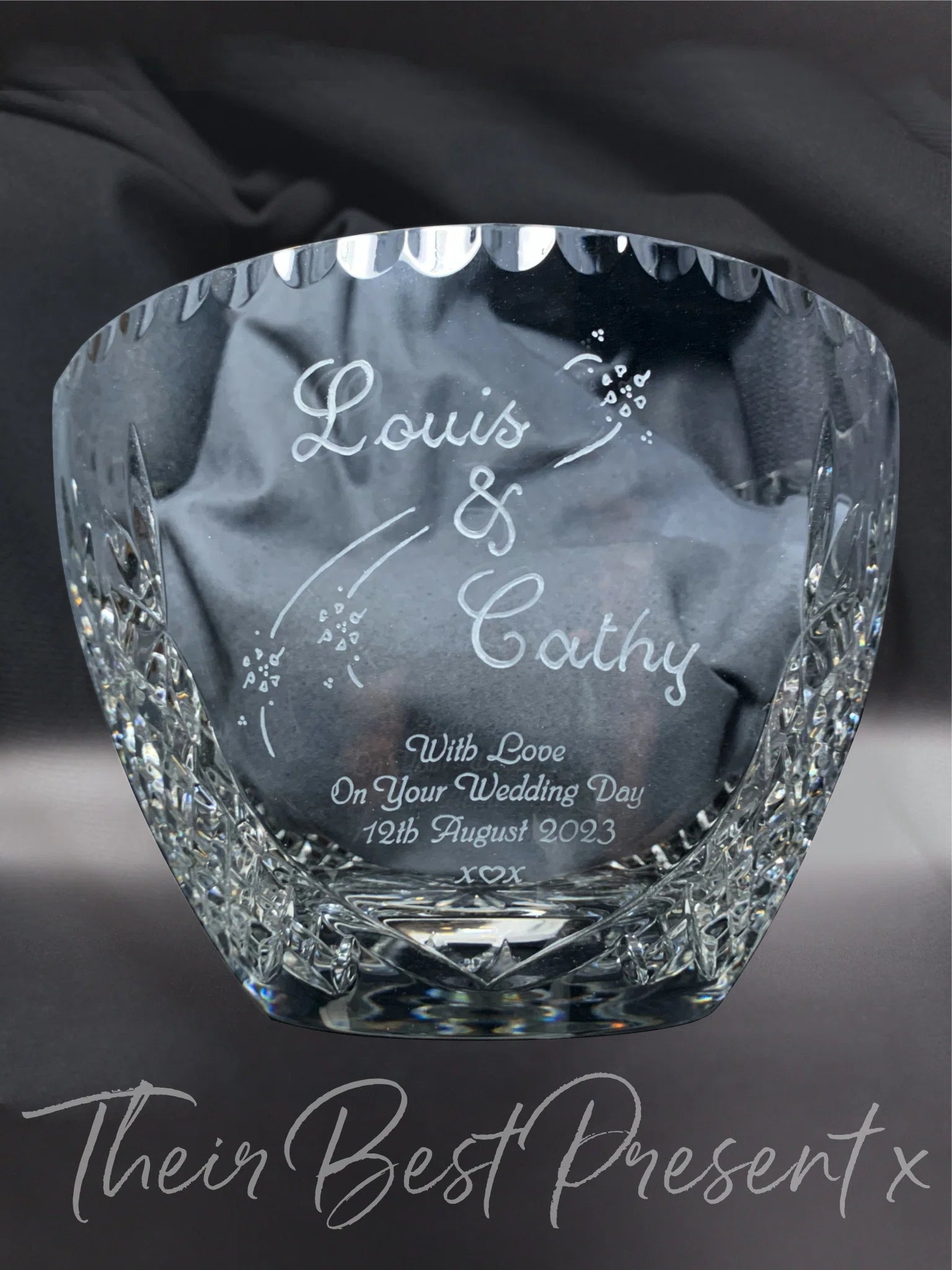 A hand-engraved and sandblasted lead crystal fruit bowl featuring "Louis & Cathy" with stylized scroll and flowers decoration, and the inscription "With Love On Your Wedding Day 12th August 2023". The piece is presented on a draped grey satin background, with overlayed text "Their Best Present" under crystal bowl capturing a bespoke and elegant gift.