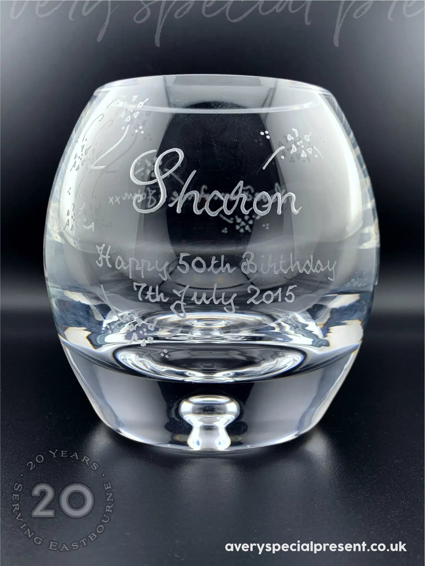 A handmade and hand-engraved candle bowl with a thick base and a transparent bubble. The bowl is inscribed with "Sharon Happy 50th Birthday 7th July 2015," featuring decorative elements and flourishes around the text. The bowl is placed on a dark surface that reflects its glassy contour and the meticulous details of the hand engraving.