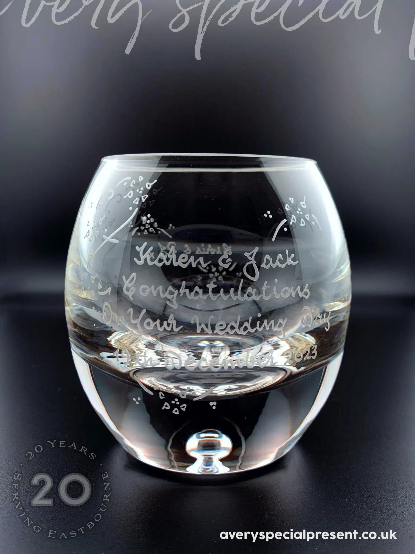 A handmade and hand-engraved candle bowl with a thick base and a transparent bubble. The bowl's front is inscribed with "Karen & Jack Congratulations On Your Wedding Day 15th December 2023." The delicate engraving includes decorative elements around the text, adding a personalized and elegant touch. The bowl sits on a reflective dark surface that enhances its clear glass and the craftsmanship of the hand engraving.