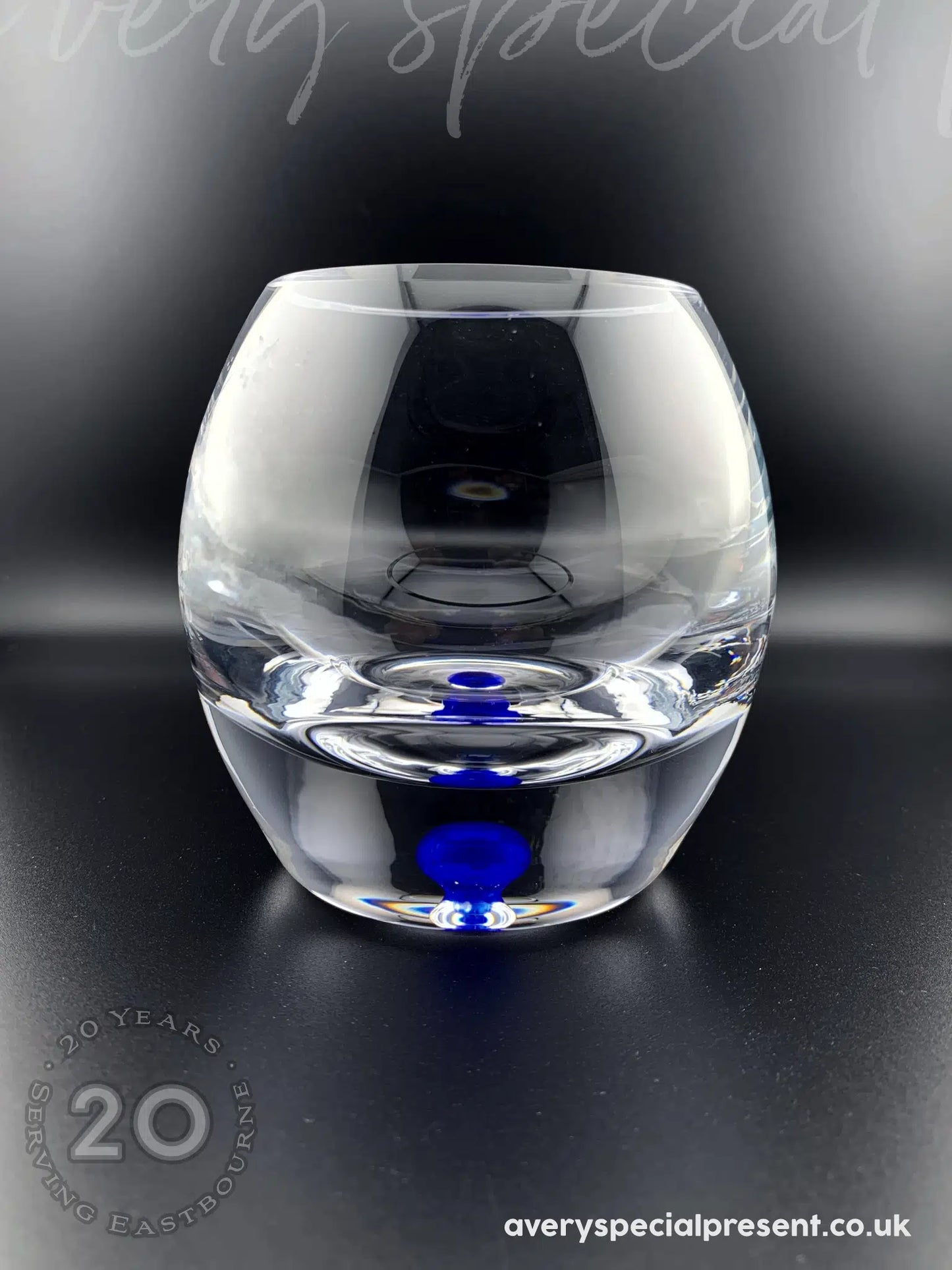 A round bubble base candle bowl with a distinctive rich blue translucent bubbles within the base. The glass's transparency creates a mesmerizing effect, showcasing the blue orb with its reflections which appear to float at different levels. The bowl is situated on a dark surface that reflects both the glass and the vibrant blue accents, highlighting the object's elegant and modern aesthetic.