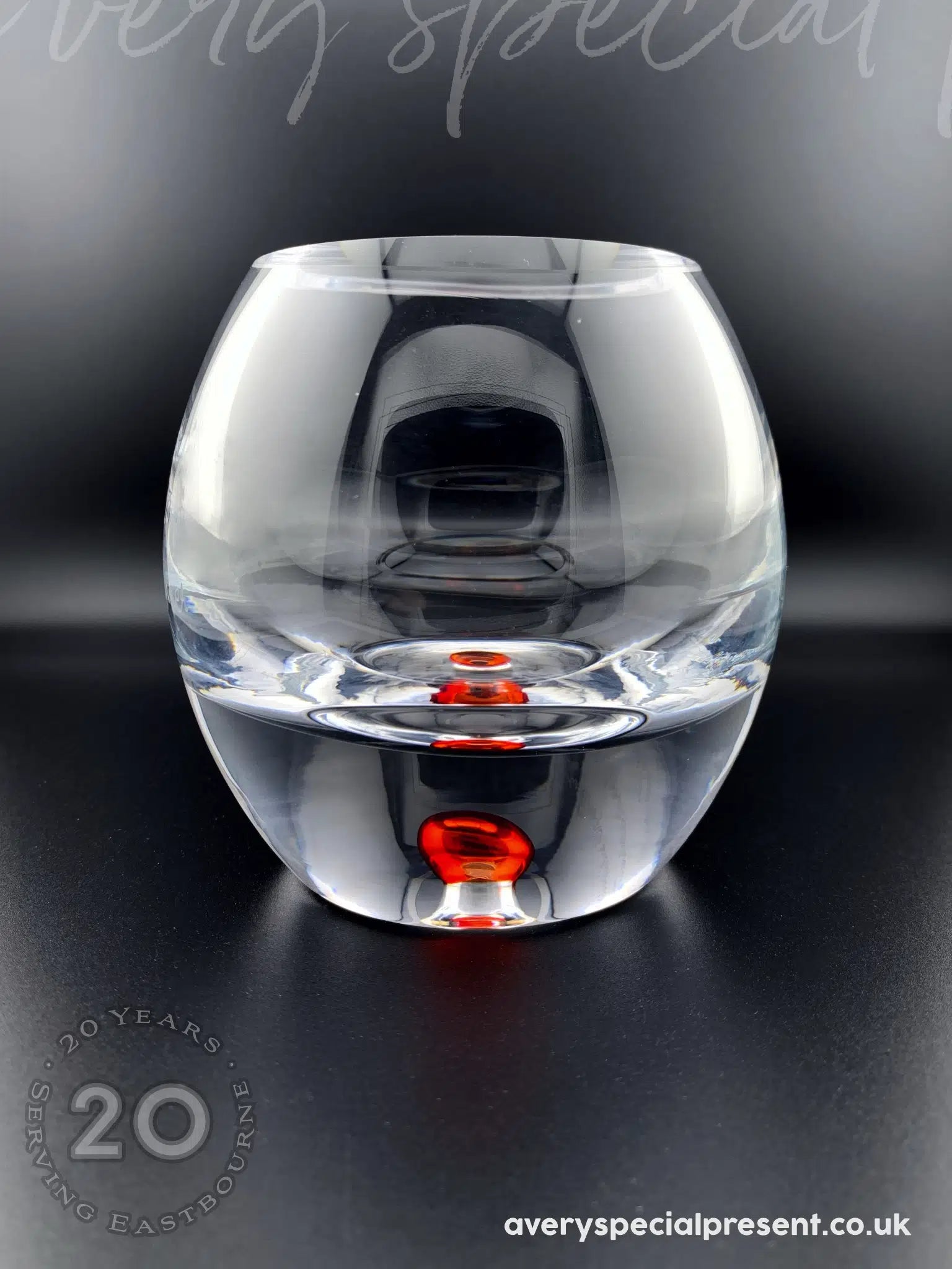 The image depicts a clear, round bubble base candle bowl with a unique design that includes a translucent crimson red bubble within the base. The transparent glass beautifully showcases the floating appearance of the red reflective elements. This piece is placed against a dark, reflective surface that accentuates its clarity and the striking colour contrast, making it a visually captivating object.