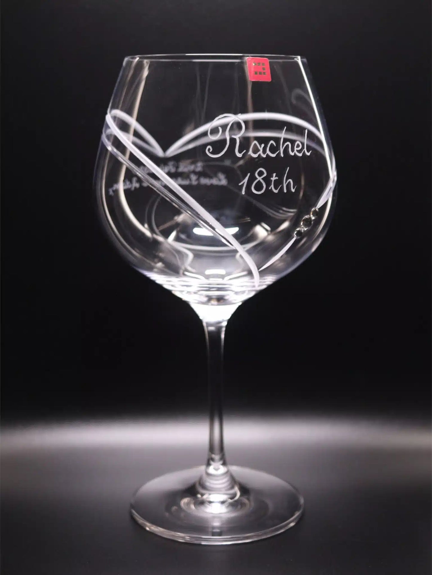 Gin glass with a long stem and a broad bowl. It features a personalized hand engraving "Rachel 18th" in a cursive hand-engraved script that sits in a large heart that frames. The heart begins on the front, finishes at the back of the glass, and has 3 Swarovski crystals. The glass is set against a dark backdrop which enhances the clarity of the glass and the detail of the engraving. Side View.