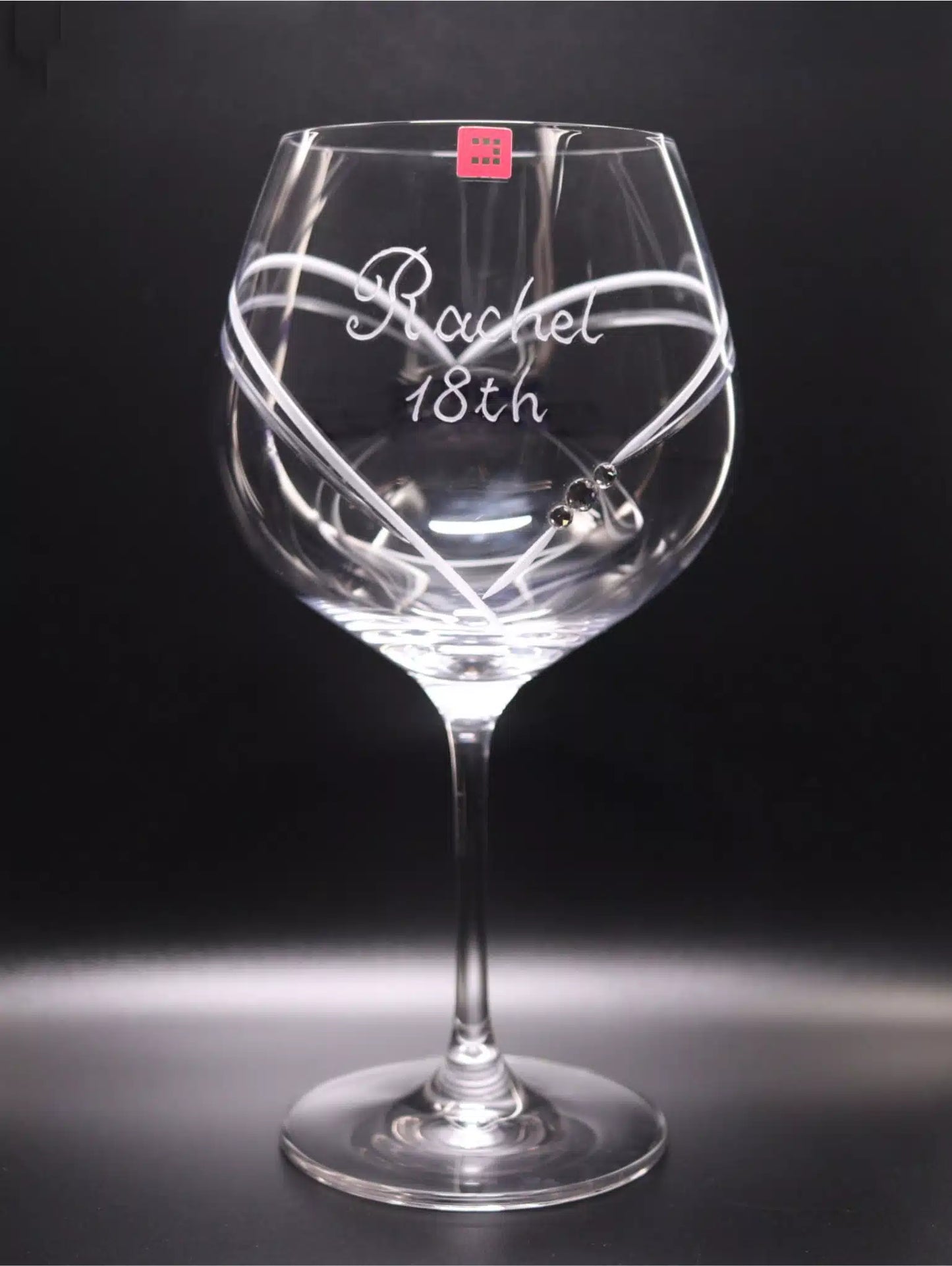 Gin glass with a long stem and a broad bowl. It features a personalized hand engraving "Rachel 18th" in a cursive hand-engraved script that sits in a large heart that frames. The heart begins on the front, finishes at the back of the glass, and has 3 Swarovski crystals. The glass is set against a dark backdrop which enhances the clarity of the glass and the detail of the engraving. Front view.