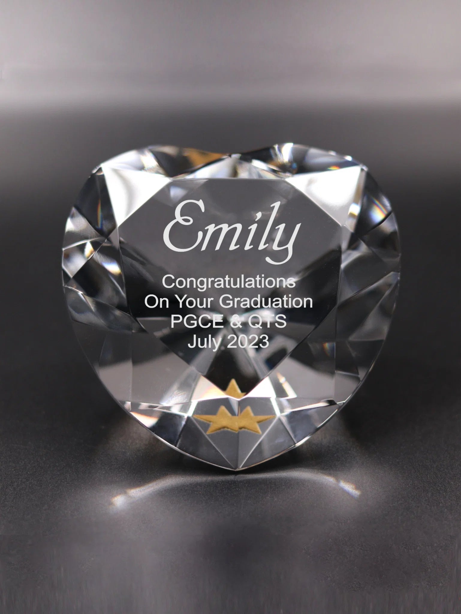 The image shows a heart-shaped, faceted crystal with an engraved message. The engraving reads "Emily Congratulations On Your Graduation PGCE & QTS July 2023" in a clean and elegant font. Beneath the text, there is a single gold star, which adds a decorative touch to the message. The background is blurred with a gradient from light to dark, which highlights the crystal's facets and the engraved text.