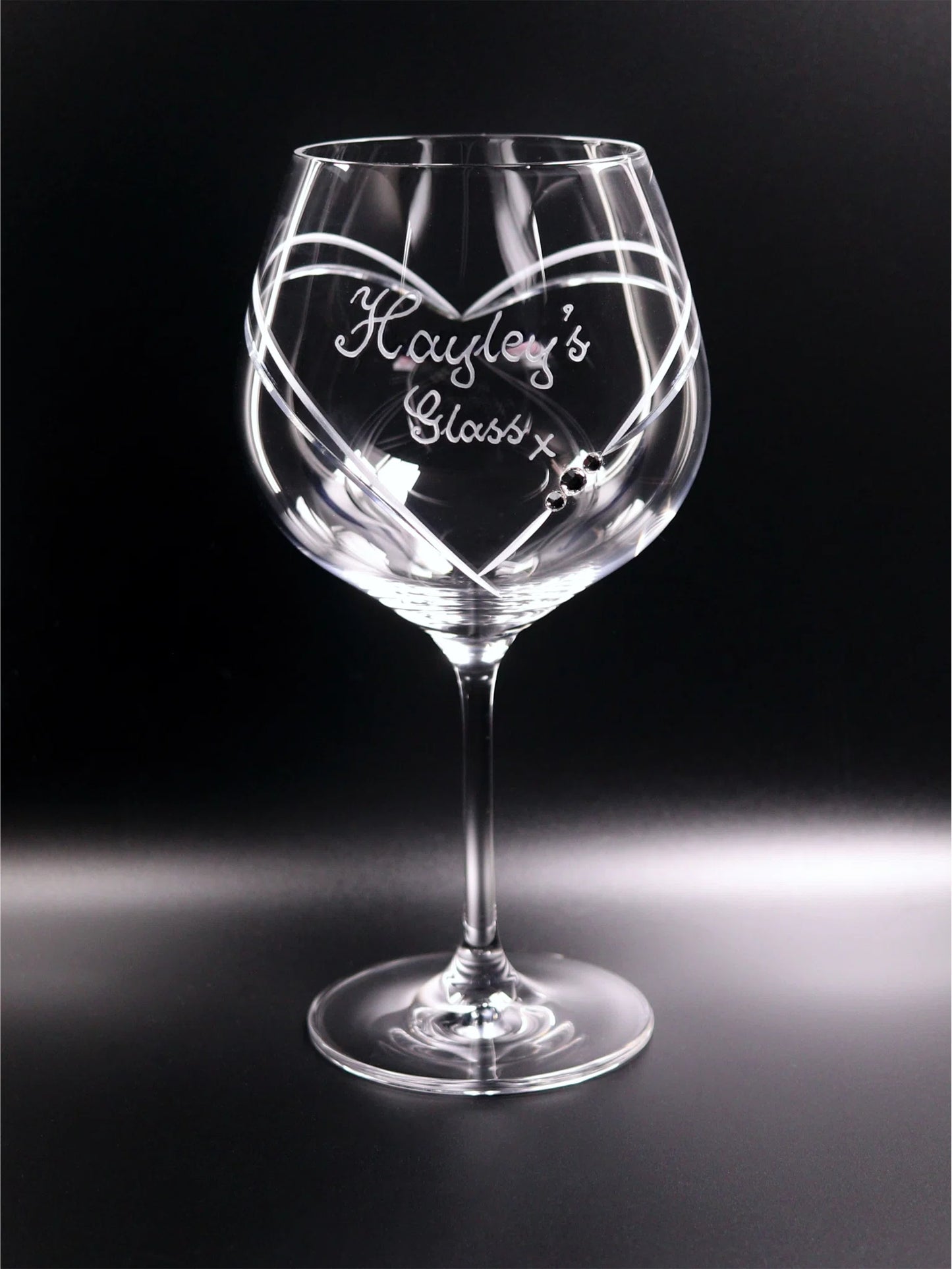 The image displays an elegant gin glass with a long stem and a broad bowl. It features a personalized hand-engraving that reads "Hayley’s Glass" with a kiss in a cursive hand-engraved script that sits in a large heart that frames. The heart begins on the front, finishes at the back of the glass, and has 3 Swarovski crystals. The glass is set against a dark backdrop which enhances the clarity of the glass and the detail of the engraving.