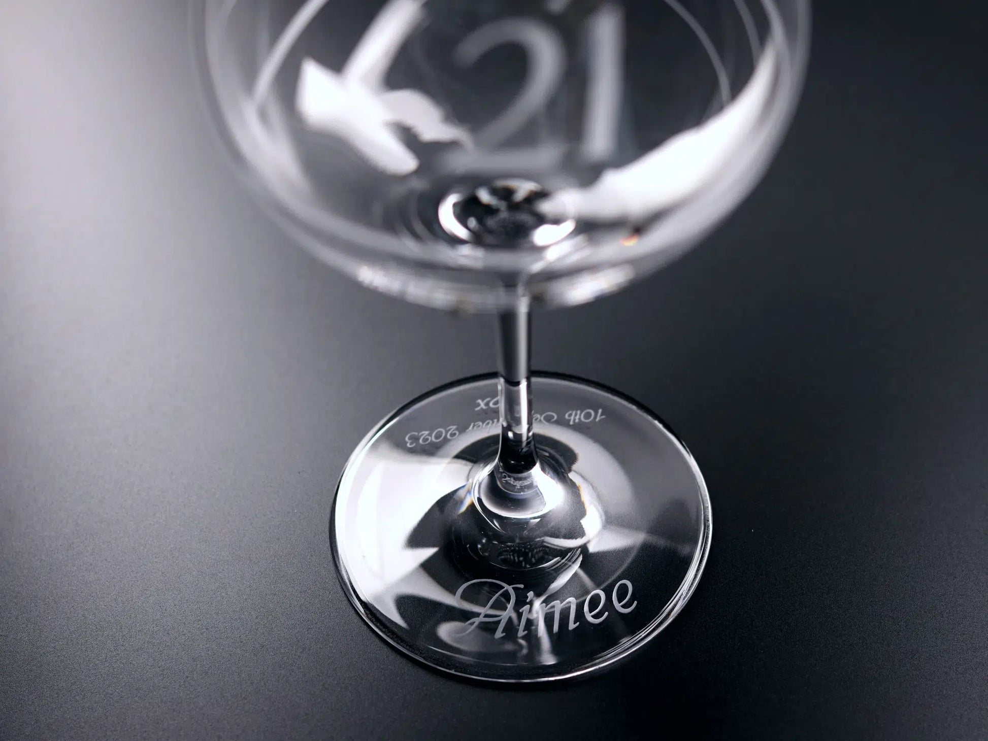 This top-down perspective photograph of a gin glass highlights its base, where the name "Aimee" elegantly arcs across the front. On the opposing side, "10th September 2023,” followed by "x♡x," is meticulously etched. The off-centre framing accentuates the glass's curves and draws the viewer's gaze across the glass's surface. Dominating the foreground is a soft blurred "21," providing an artistic juxtaposition to the crisp engraving.