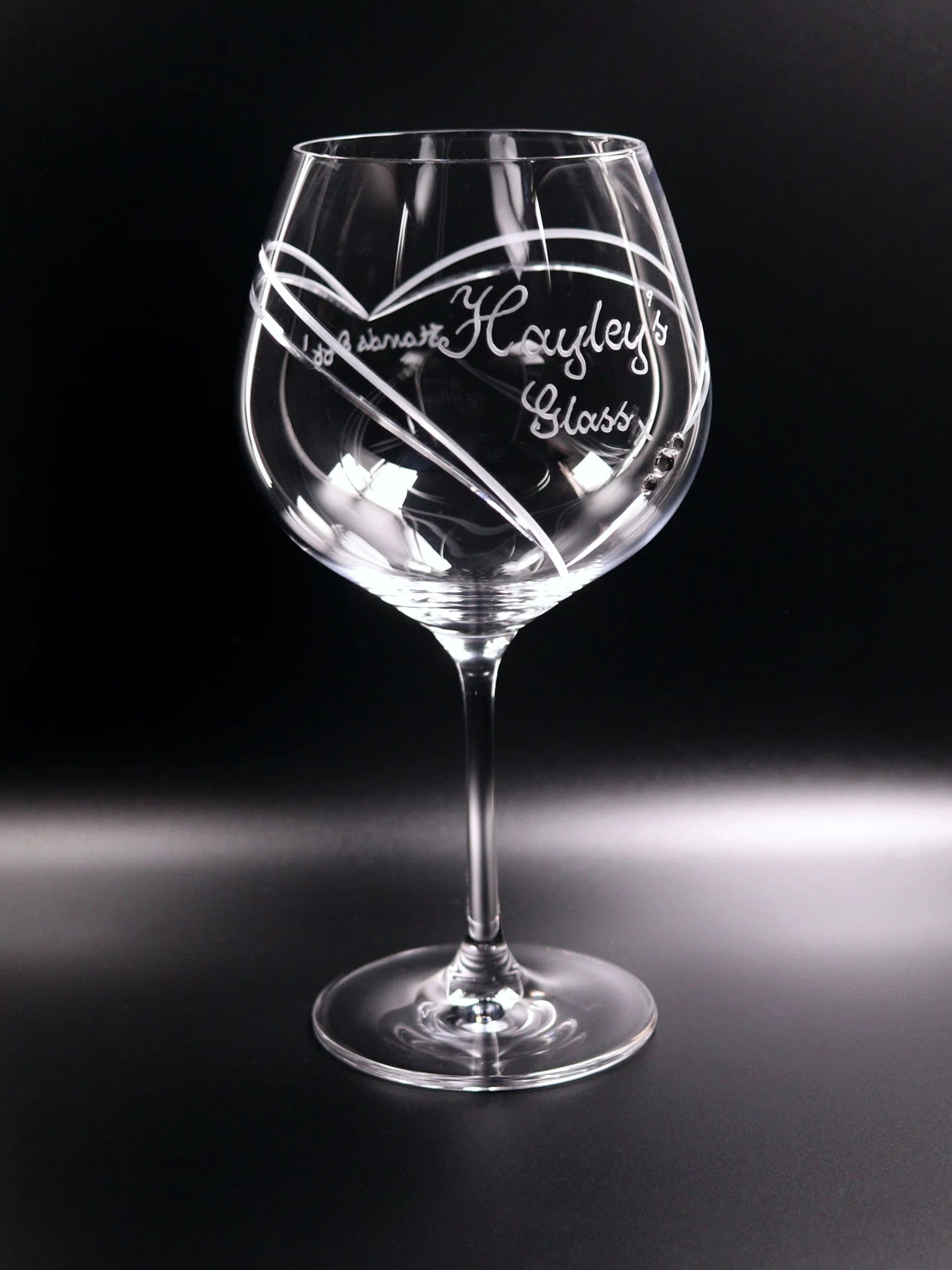 An elegantly crafted gin glass with a long stem and expansive bowl, personalized with hand-engraved cursive script reading "Hayley’s Glass," punctuated with a kiss mark. The glass, slightly turned, reveals a large, artfully framed heart design that wraps from the front to the back, embellished with three Swarovski crystals for a touch of sparkle. Set against a dark background, the clear glass and intricate engraving are prominently showcased, highlighting the glass's exquisite detail and bespoke design.