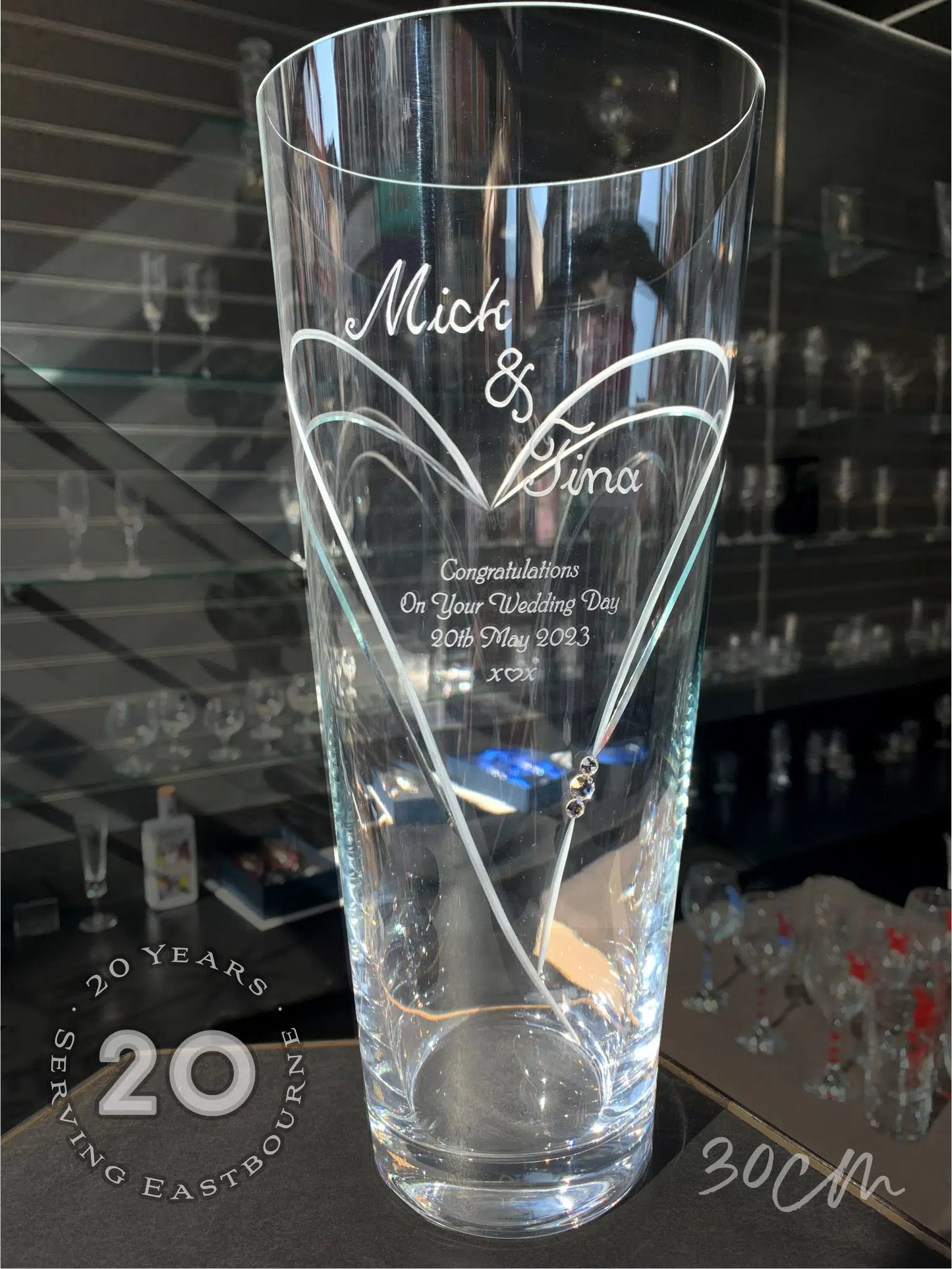 A tall, 30cm Diamante Conical Vase with Heart Shaped Cutting with 3 Swarovski® Crystals displaying "Mick & Tina Congratulations On Your Wedding Day 20th May 2023 x ♡ x", with a stylized design and two embedded crystals. Text overlay commemorates 20 years serving Eastbourne, showcased in a well-lit display.