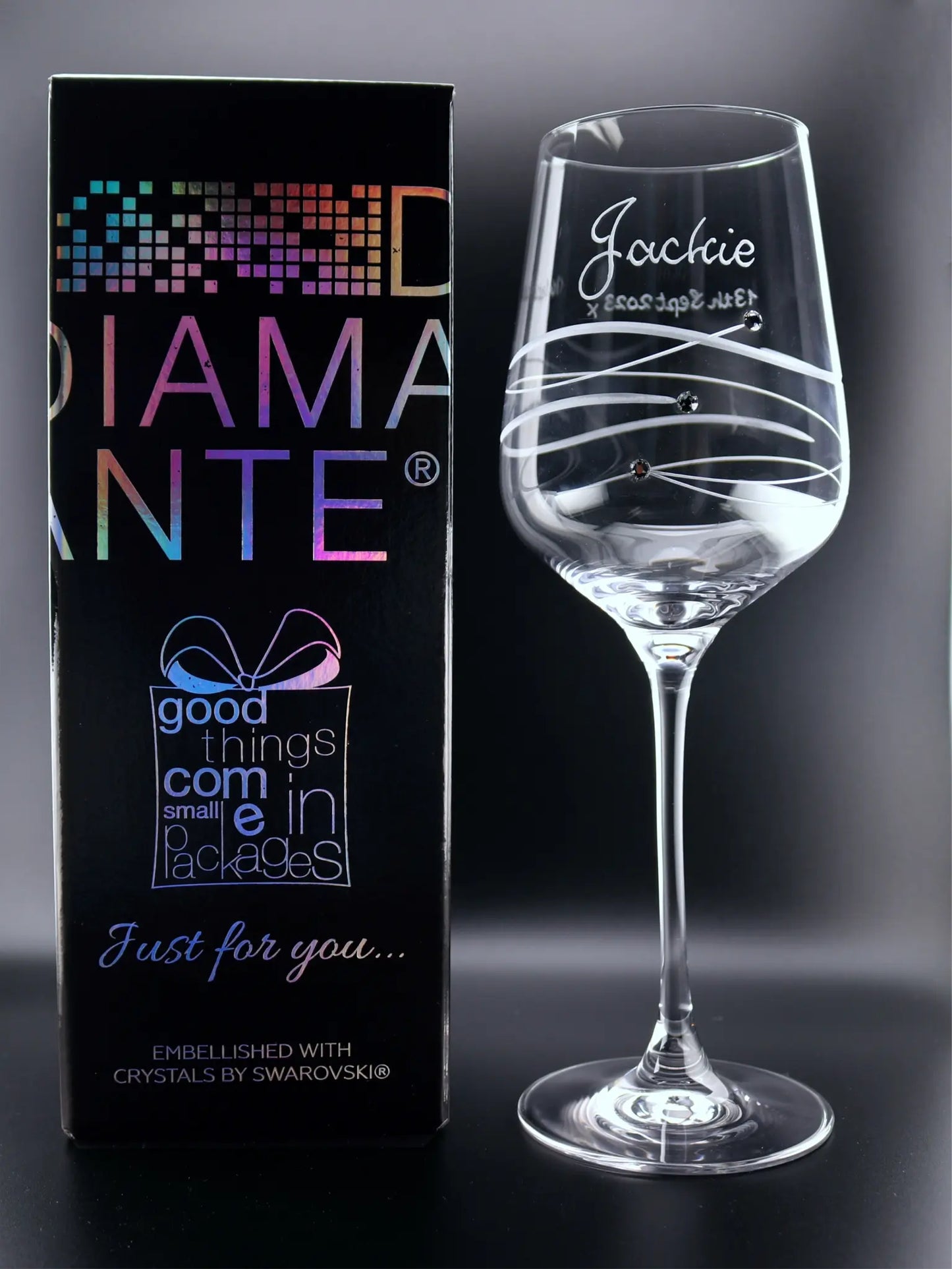 A personalized wine glass with "Jackie" elegantly hand-engraved on the face, alongside swirling lines cut into the glass and three Swarovski crystals. The glass is showcased next to its packaging, a DIAMANTE box with holographic text reading "good things come in small packages Just for you…" and the promise of Swarovski crystal embellishment.