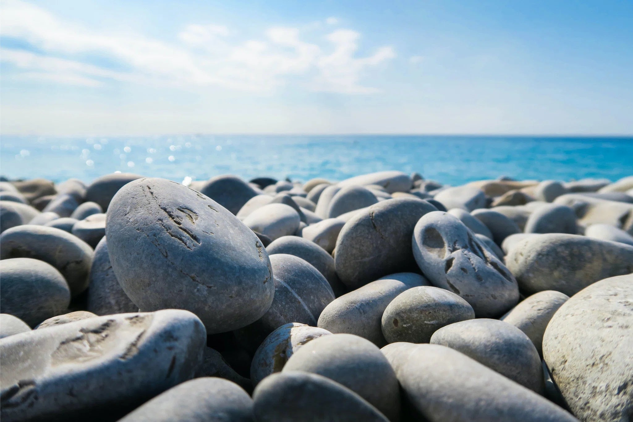 Smooth, rounded pebbles cover a beach, their pale gray surfaces basking in sunlight. The horizon reveals a tranquil blue sea meeting a clear sky, while sunbeams dance over the gentle waves, adding sparkle to this serene coastal scene.