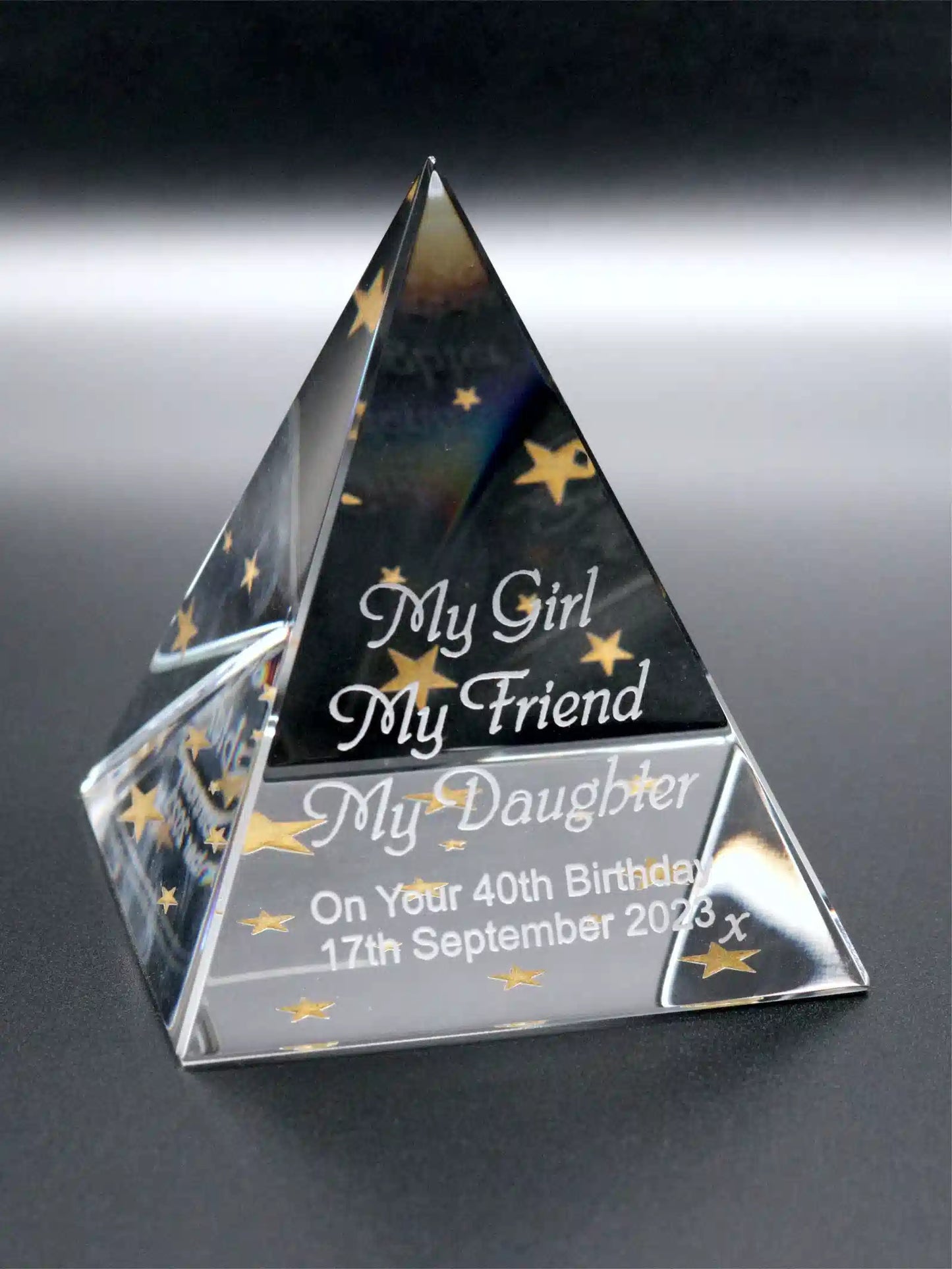 A crystal-clear glass pyramid award with a reflective surface stands at an angle, capturing the light. It's intricately engraved with the heartfelt inscription "My Girl, My Friend, My Daughter, On your 40th Birthday, 17th September 2023 x" in a graceful script, accented by delicate stars scattered around the text, which add a touch of whimsy. The craftsmanship showcases precision in the etched lettering, giving the piece a personalized and timeless charm.