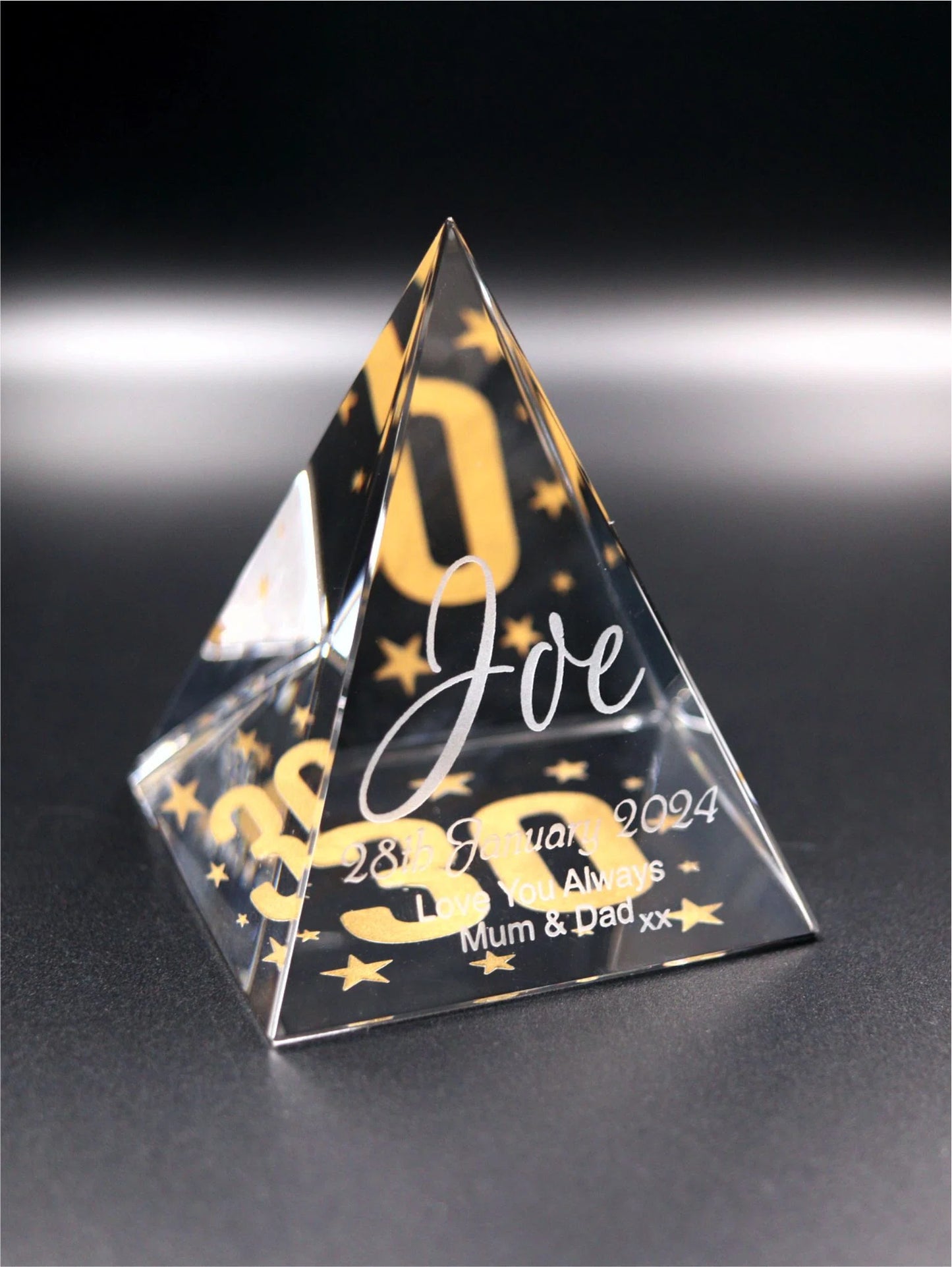 The image shows a clear glass pyramid with a personalized engraving. The engraving includes the name "Joe" in stylized script at the center, surrounded by decorative elements such as stars and what appears to be the number "30" in a larger font size. Below the name, there is a date "28th January, 2024" and a sentimental message "Love You Always Mum & Dad xx". The background is blurred, emphasizing the pyramid and its engravings.