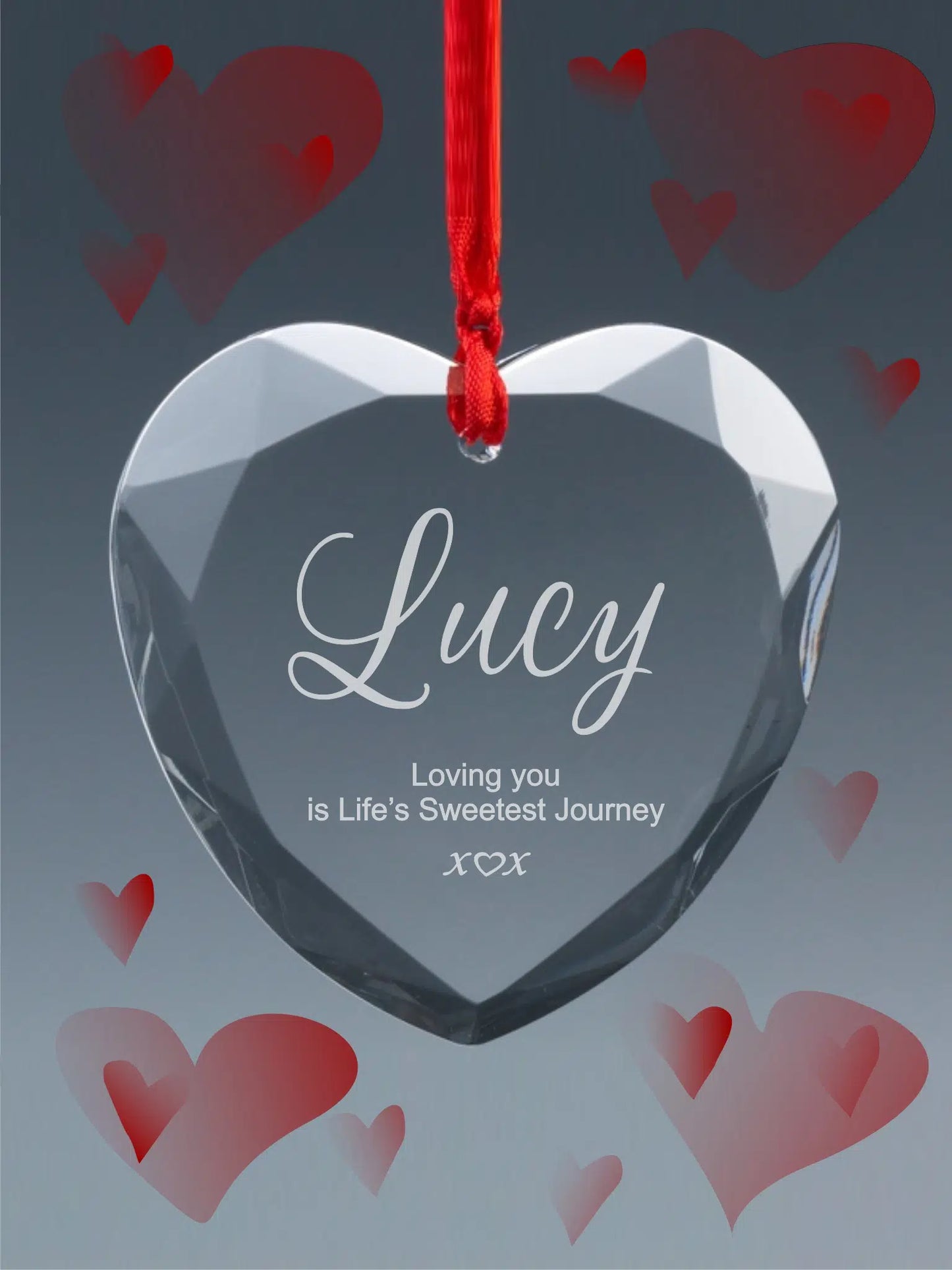 A clear heart-shaped crystal engraved with the name 'Lucy' and the phrase 'Loving you is Life's Sweetest Journey', suspended by a red ribbon, with translucent red hearts in the background.