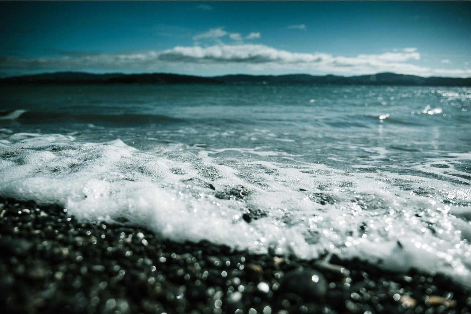 Foamy sea wave gently washing over a pebble beach with a clear blue sky and distant mountains on the horizon.